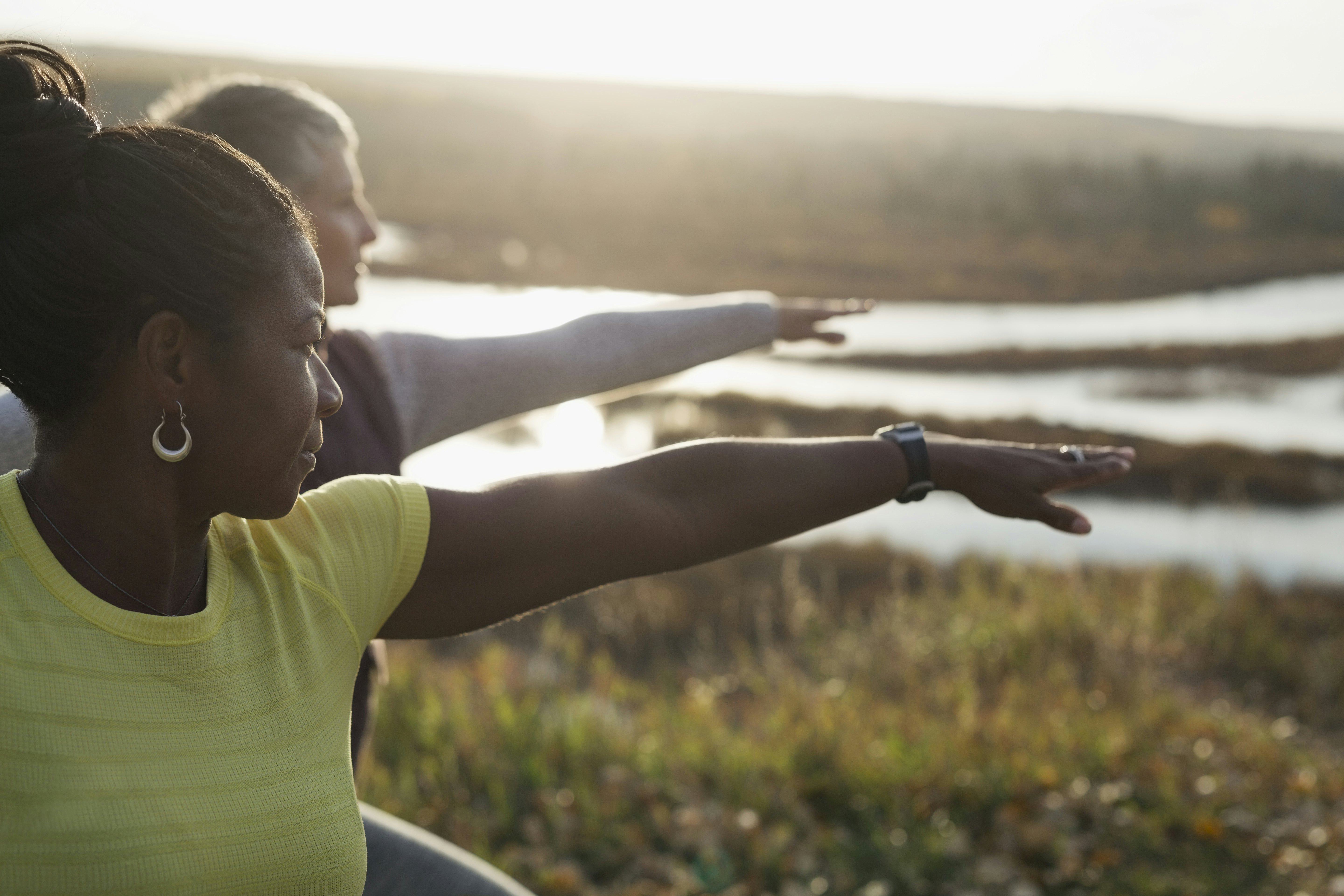 Two women practising a yoga pose (warrior 2) outdoors on a sunny afternoon.