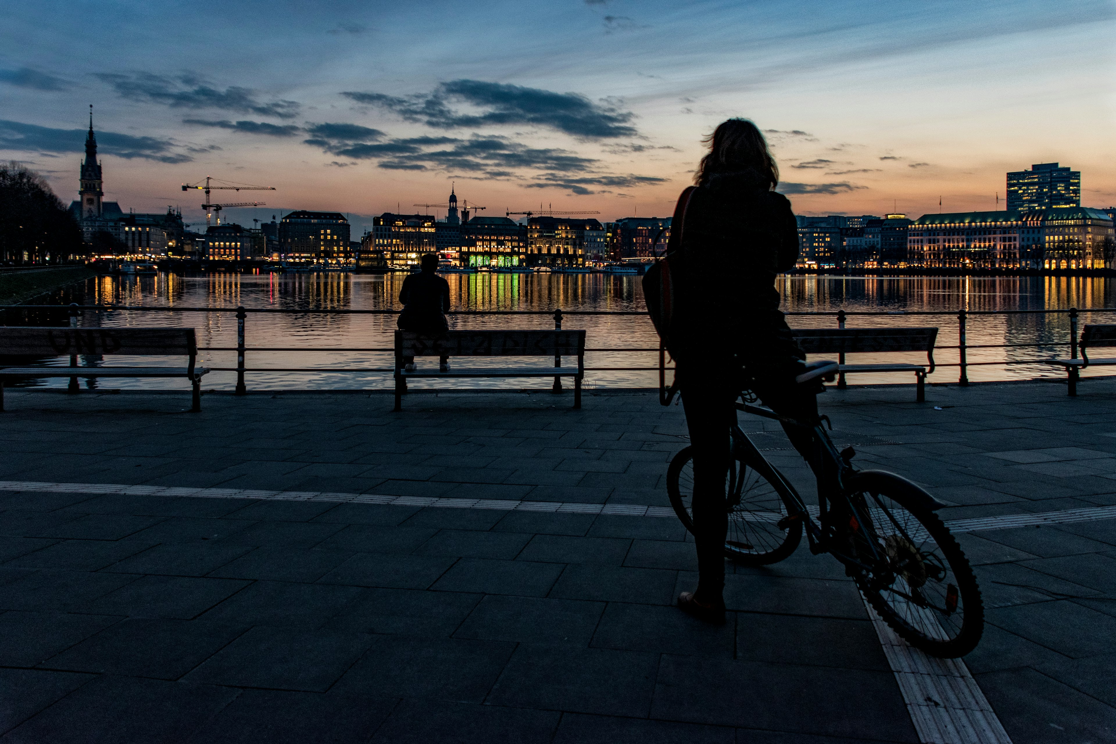 See Hamburg in a weekend by bike. A photo of a cyclist overlooking the canal at dusk. We can see one of Hamburg's famous spires silhouetted against the colourful sky, the lights from the buildings are reflected in the water.