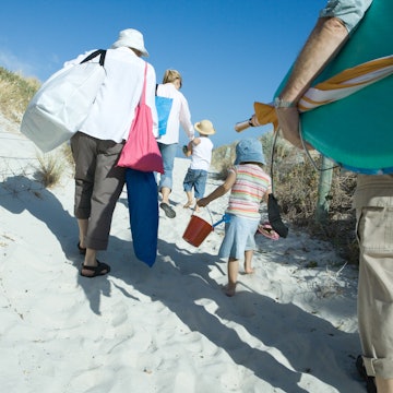 Rear view of a family group of three adults and two young children, make their way through sand dunes carrying a lot of beach items
