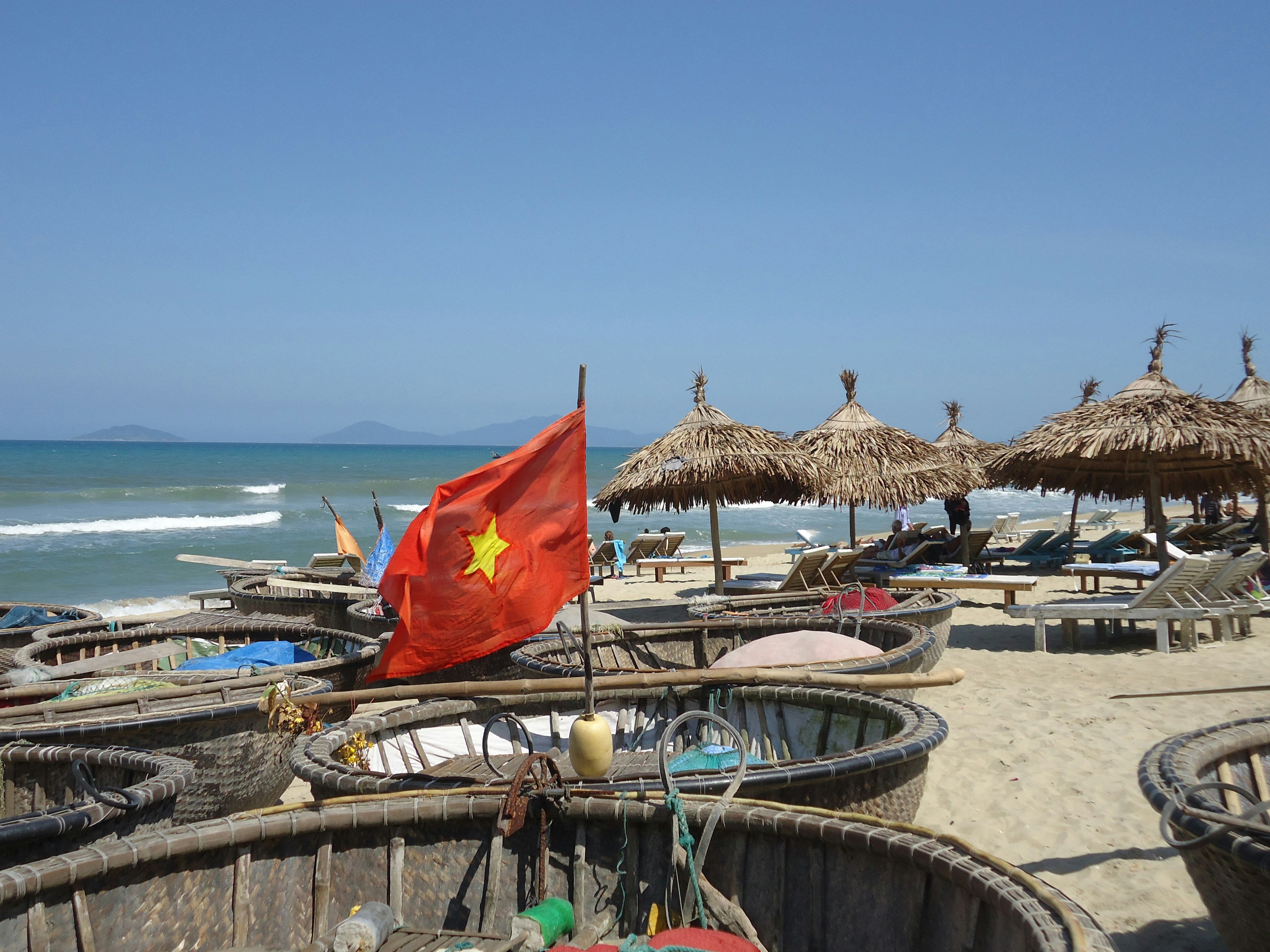 A Vietnamese flag flies above a basket boat on the beach, with some sunshades and sunloungers in the background