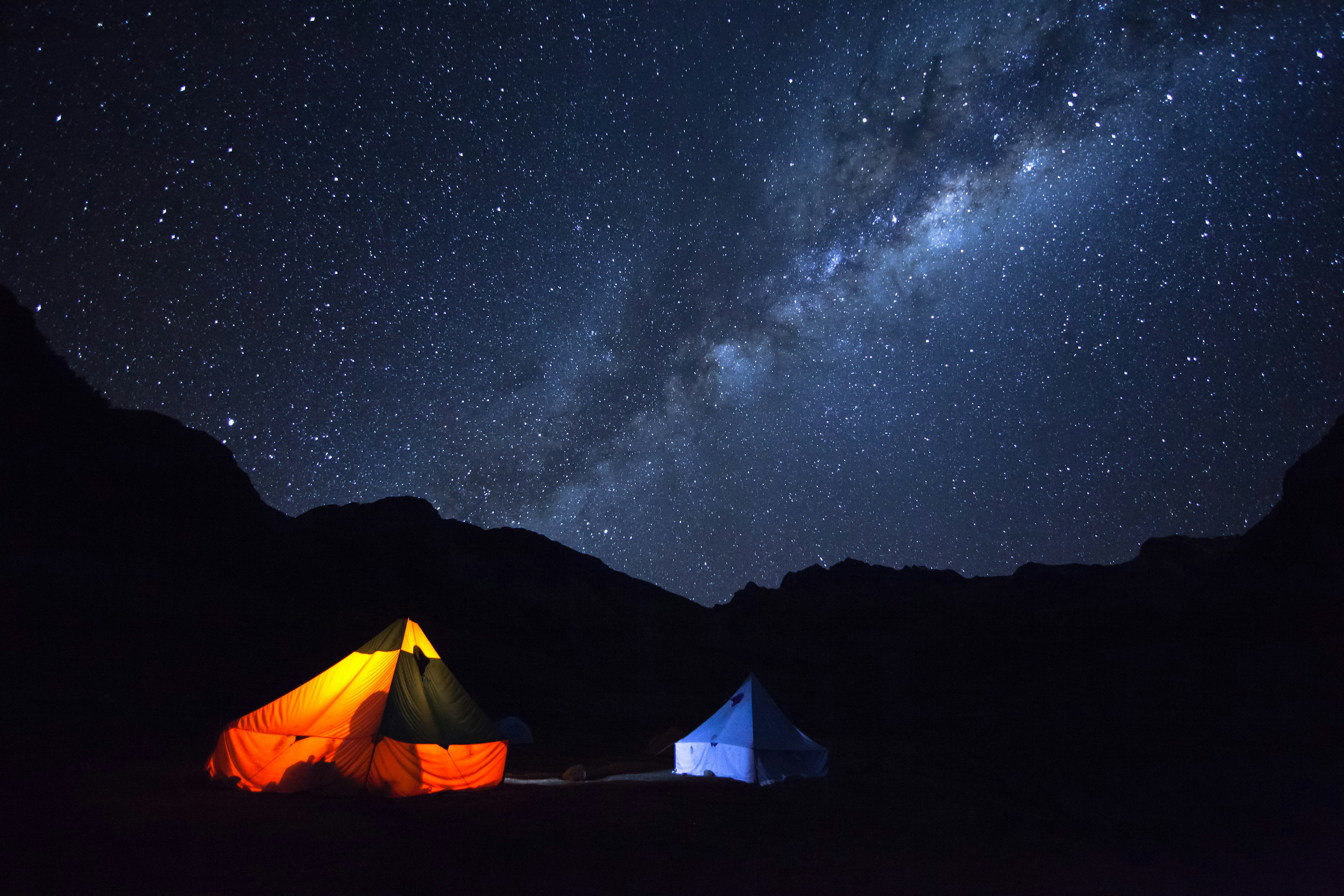 A nighttime shot of two lit-up tents in the foreground with a starry sky in the background (Milky Way visible)