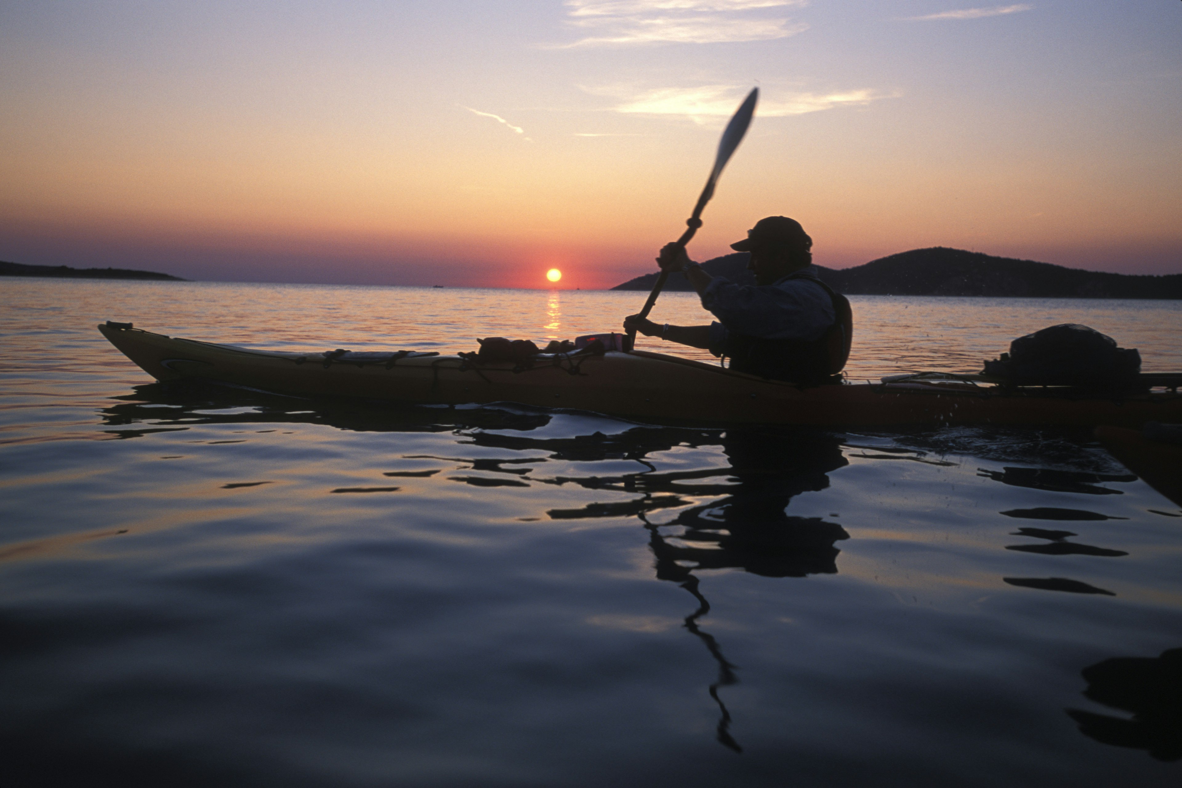 A silhouette of a man piloting a kayak in a lake amongst a serene sunset.