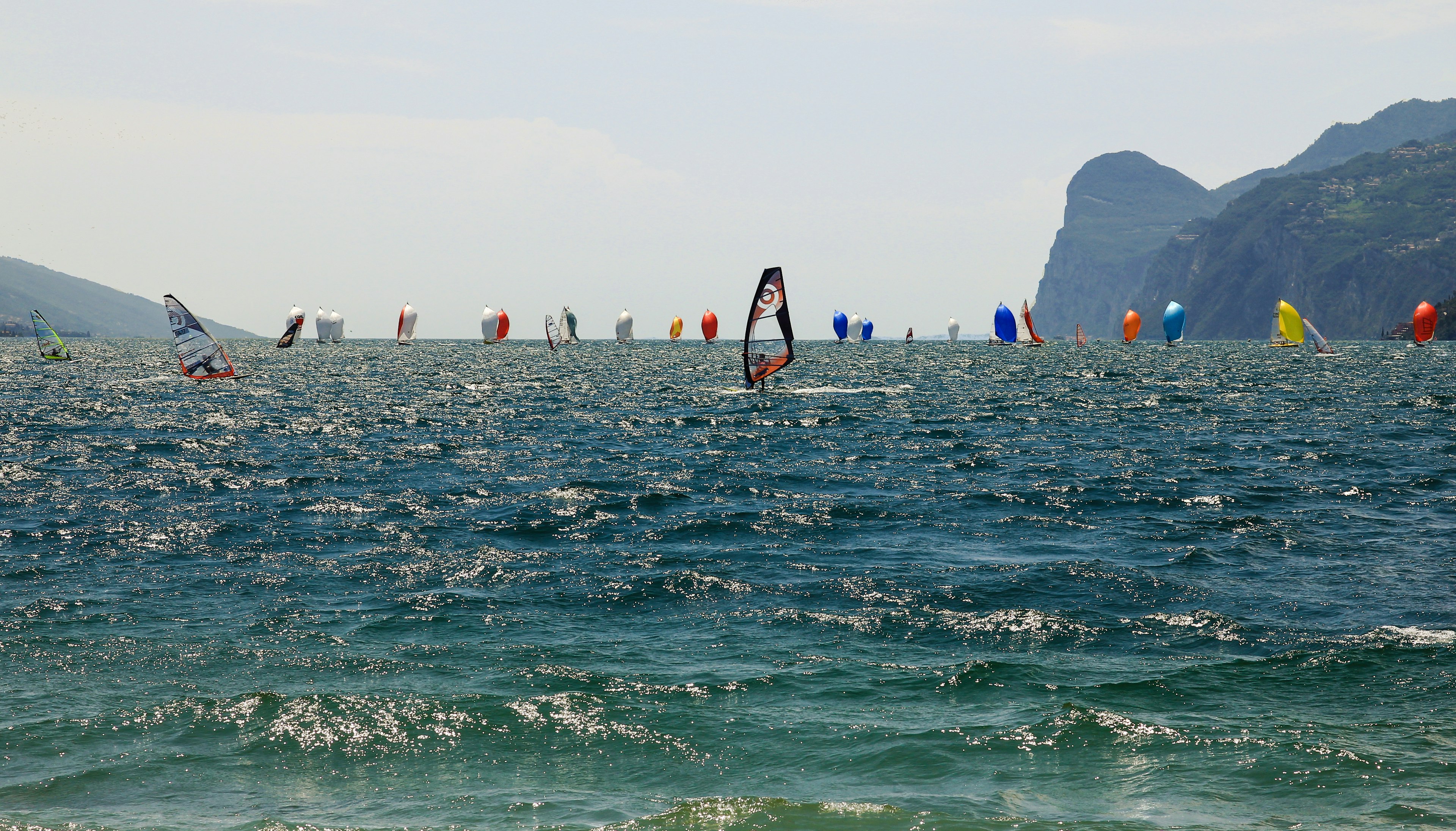 Sailing regatta in Lake Garda with plenty of boats and colored sails with a few windsurfers in the foreground.