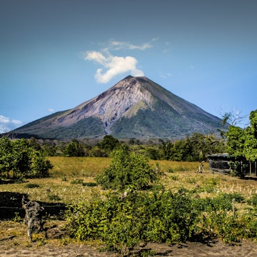 A white plume of smoke escapes from the top a mountain. In front of the mountain is a collection of low-lying shrubs with larger trees framing the sides of the image. Nicaragua travel is still an area of concern for potential visitors.