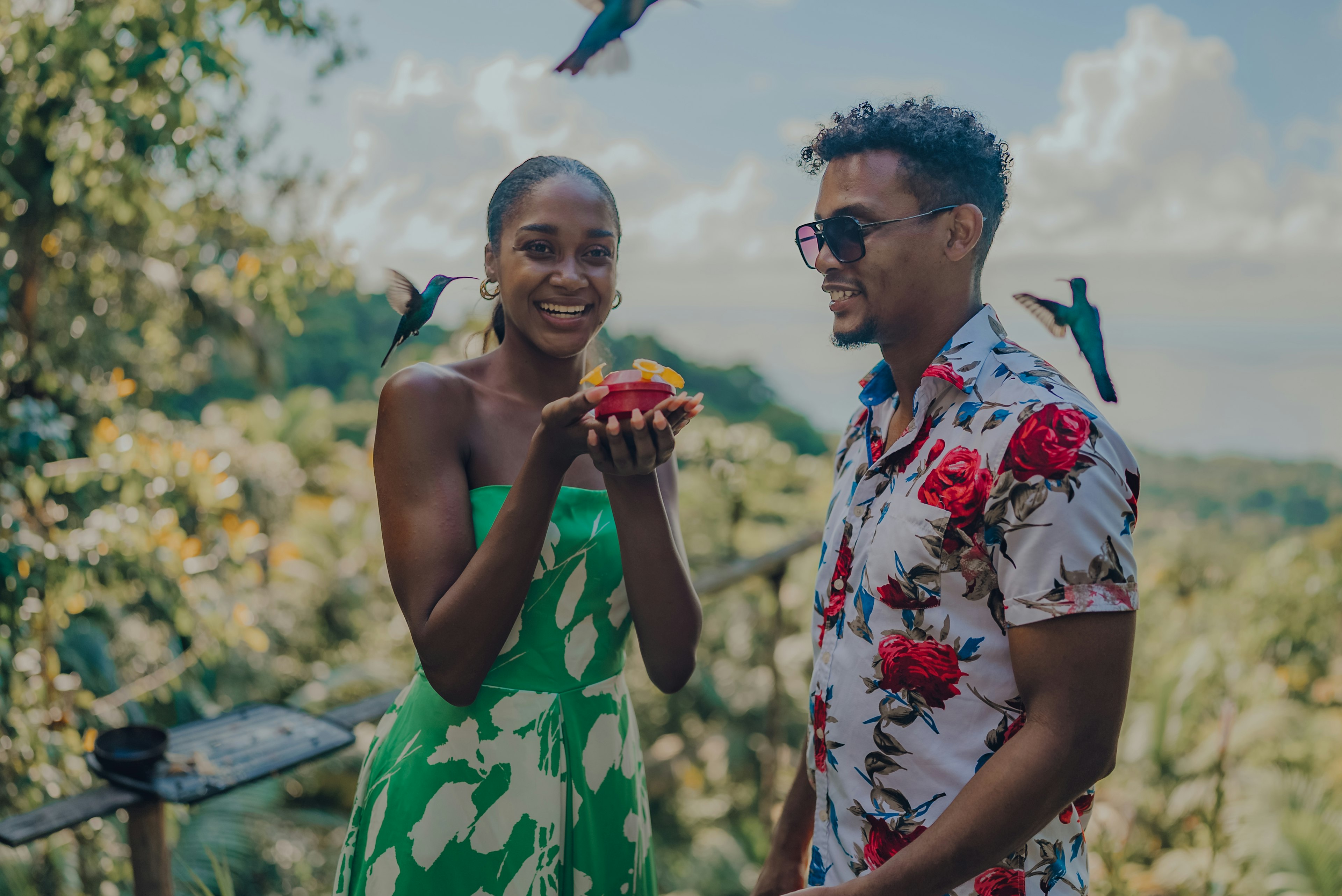 A young couple in brightly colored clothing smiles as they feed hummingbirds in Tobago, Trindad & Tobago