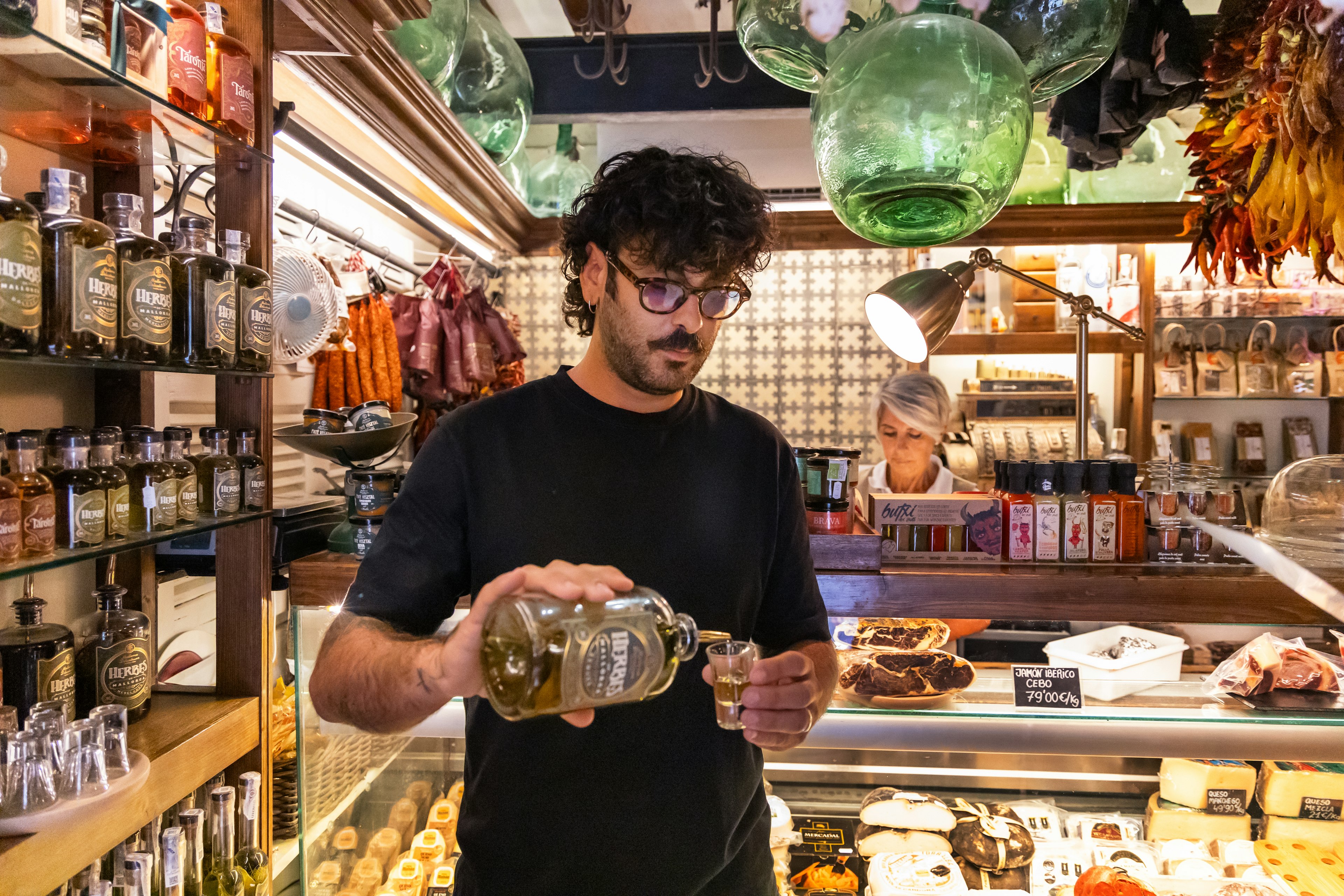 A local vendor pours a traditional Mallorcan herbal liqueur at a deli counter in Palma de Mallorca, offering visitors a taste of authentic flavors.