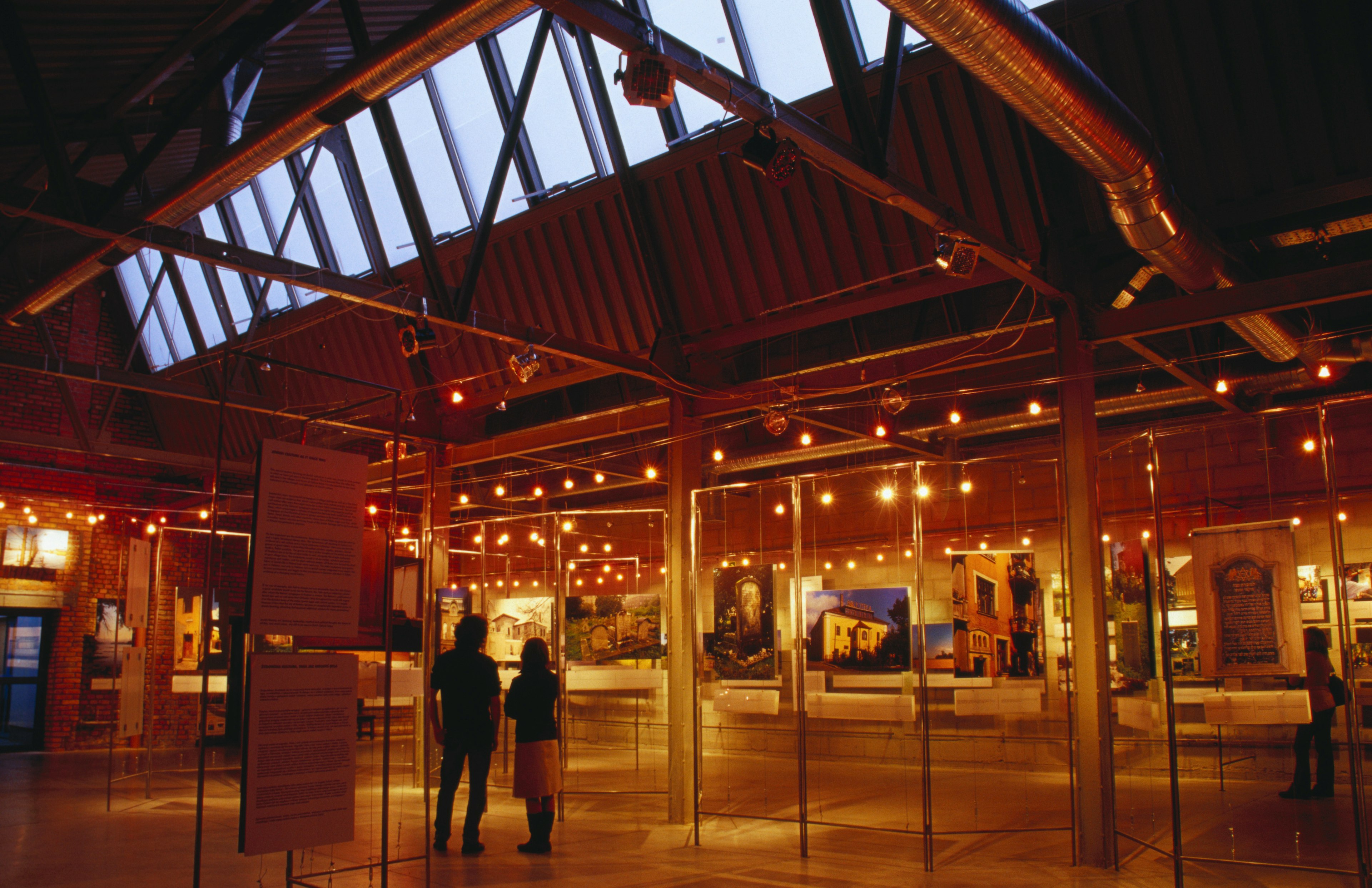 Photographic displays hang on boards around a warmly lit room. Two people are looking at one of the displays