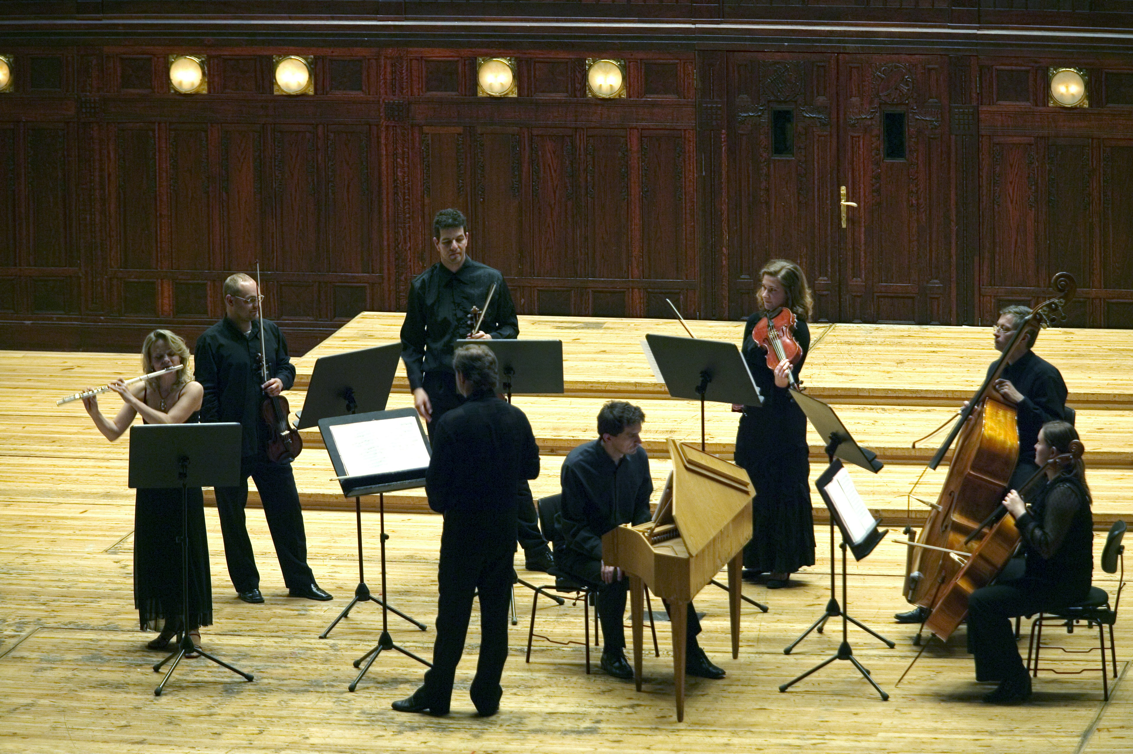 Eight classical musicians, playing flutes, cellos and violins, stand on a wooden stage