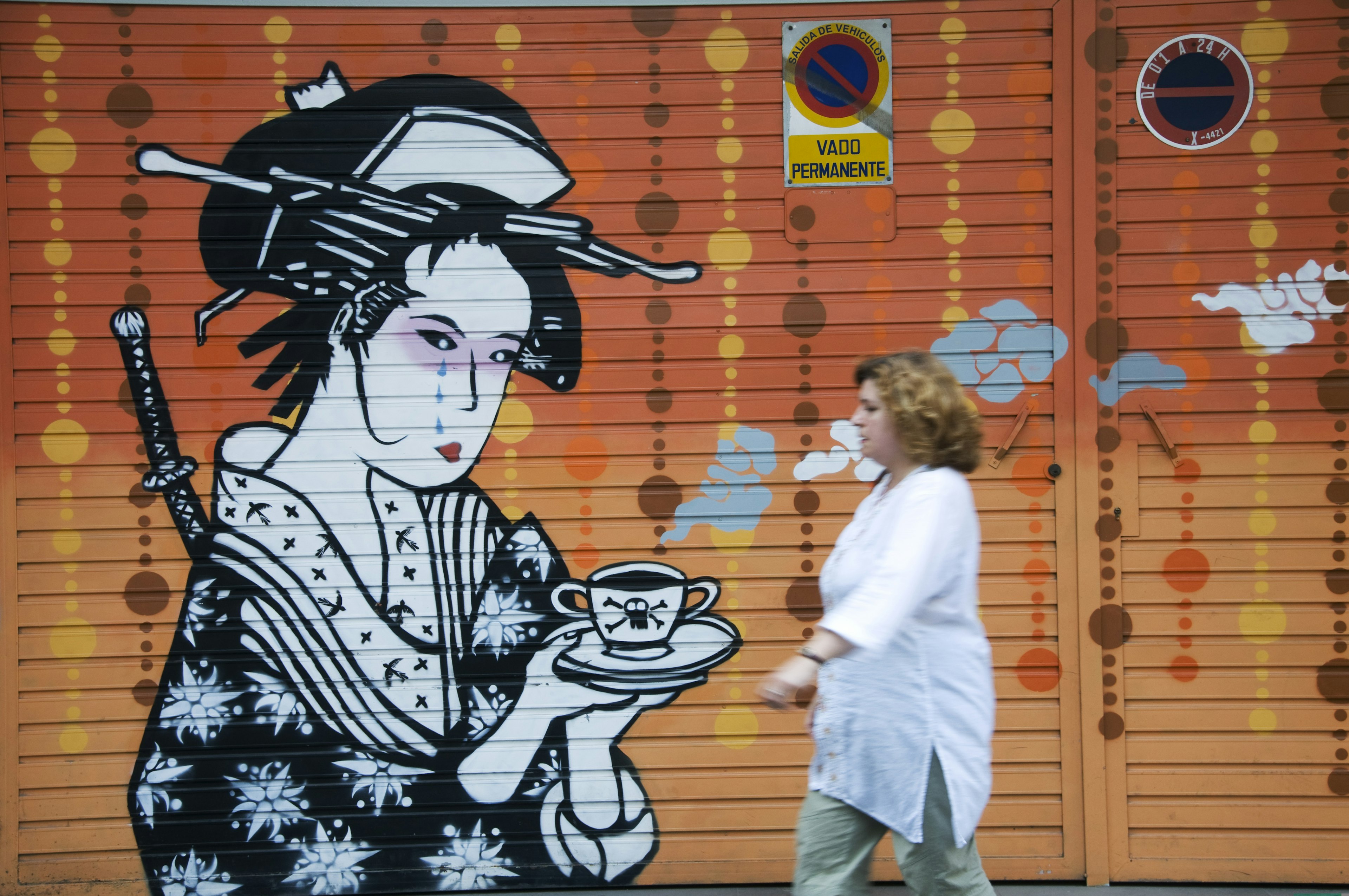 A woman walks by a painting depicting a woman in Japanese clothing on a storefront gate in the Russafa district of Valencia, Spain