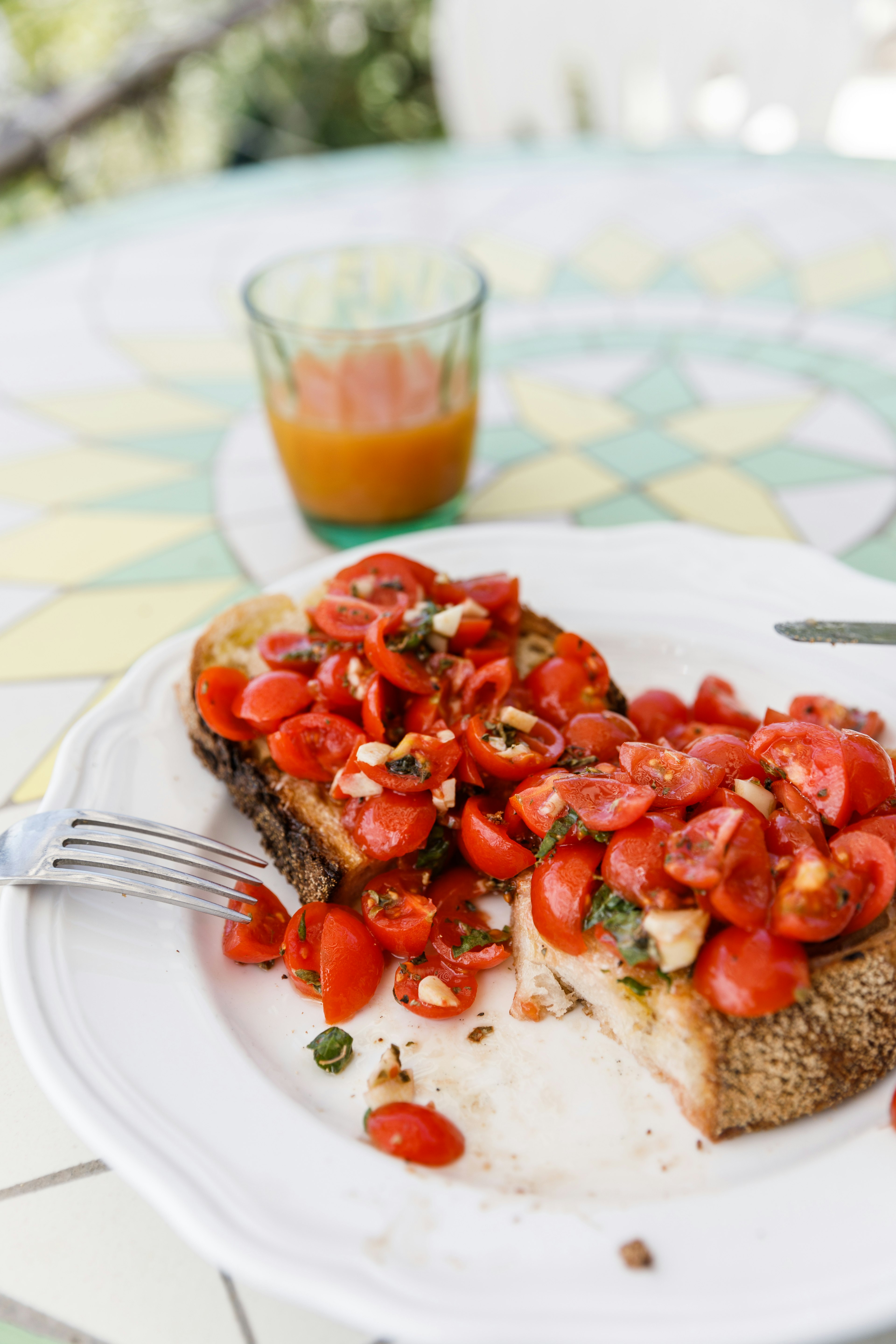 A close up shot of two slices of bruschetta. The crusty bread is piled with tomatoes, oil and garlic on a colourfully-tiled table with a glass of juice out of focus in the background.