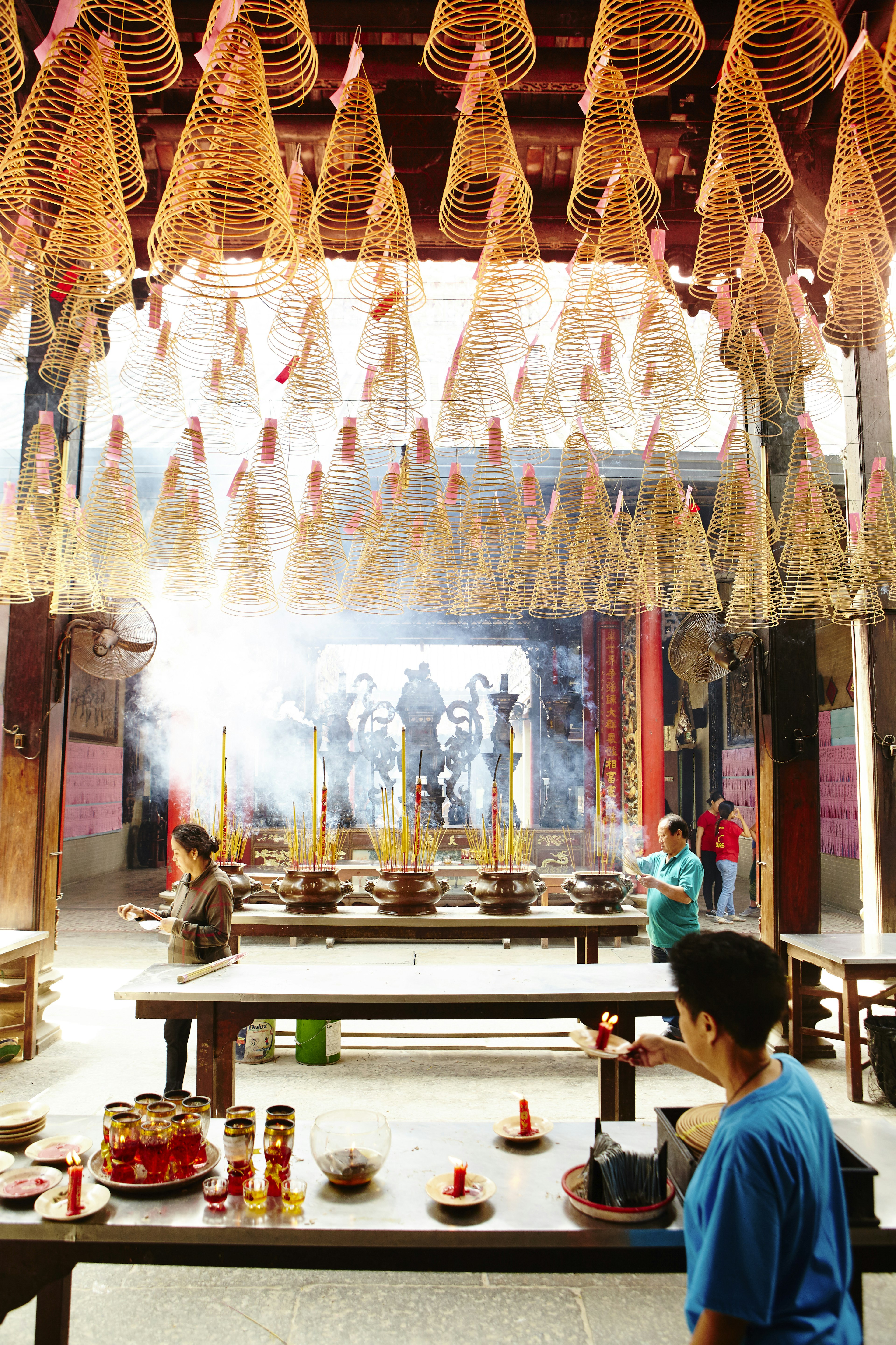 Large cone-shaped coils of incense hang from the ceiling of Thien Hau Pagoda, incense sticks give off smoke, while a visitor lights a candle below