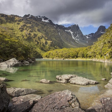 Wide image of Lake MacKenzie on New Zealand's South Island on a cloudy day, large rocks in the foreground and mountains in the background.