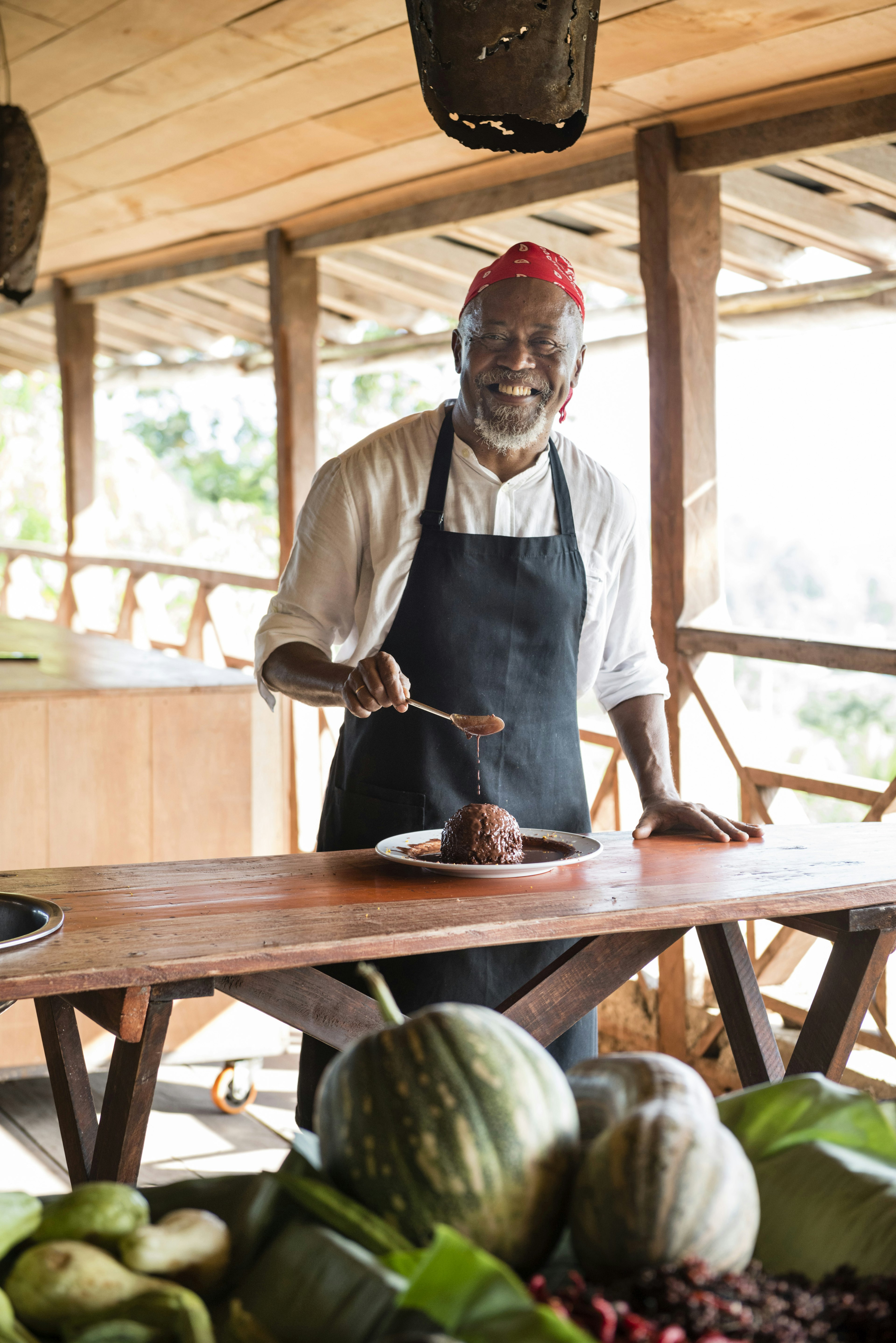 The smiling chef pours sauce over a dish with a spoon. The setting is a wooden building without walls. The plate sits on a wooden table