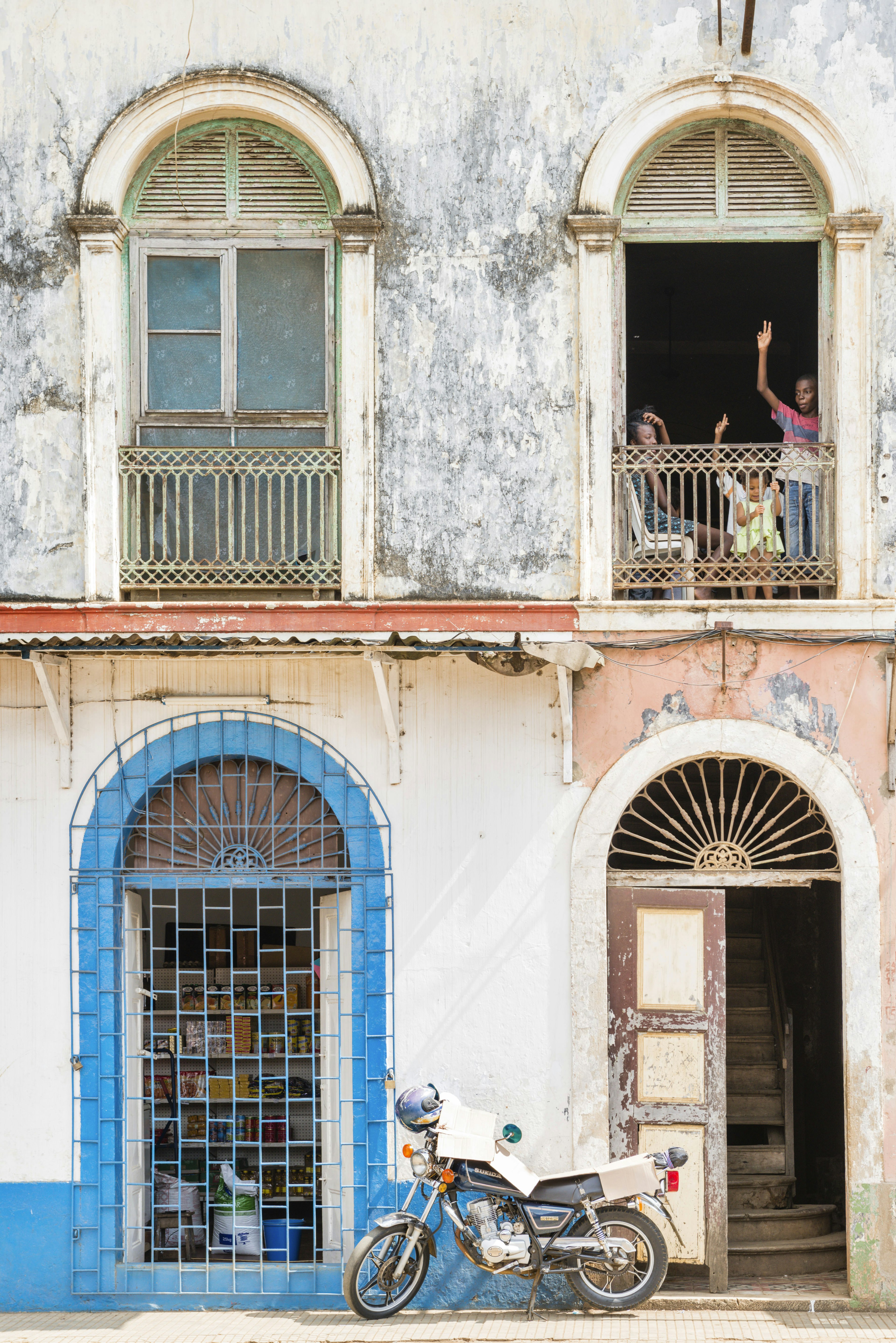 An old motorbike, with helmet sittiing on the side-view mirror stands in front of a decaying white-washed builing with dilapidated doors and windows. A family waves from one of the upper windows