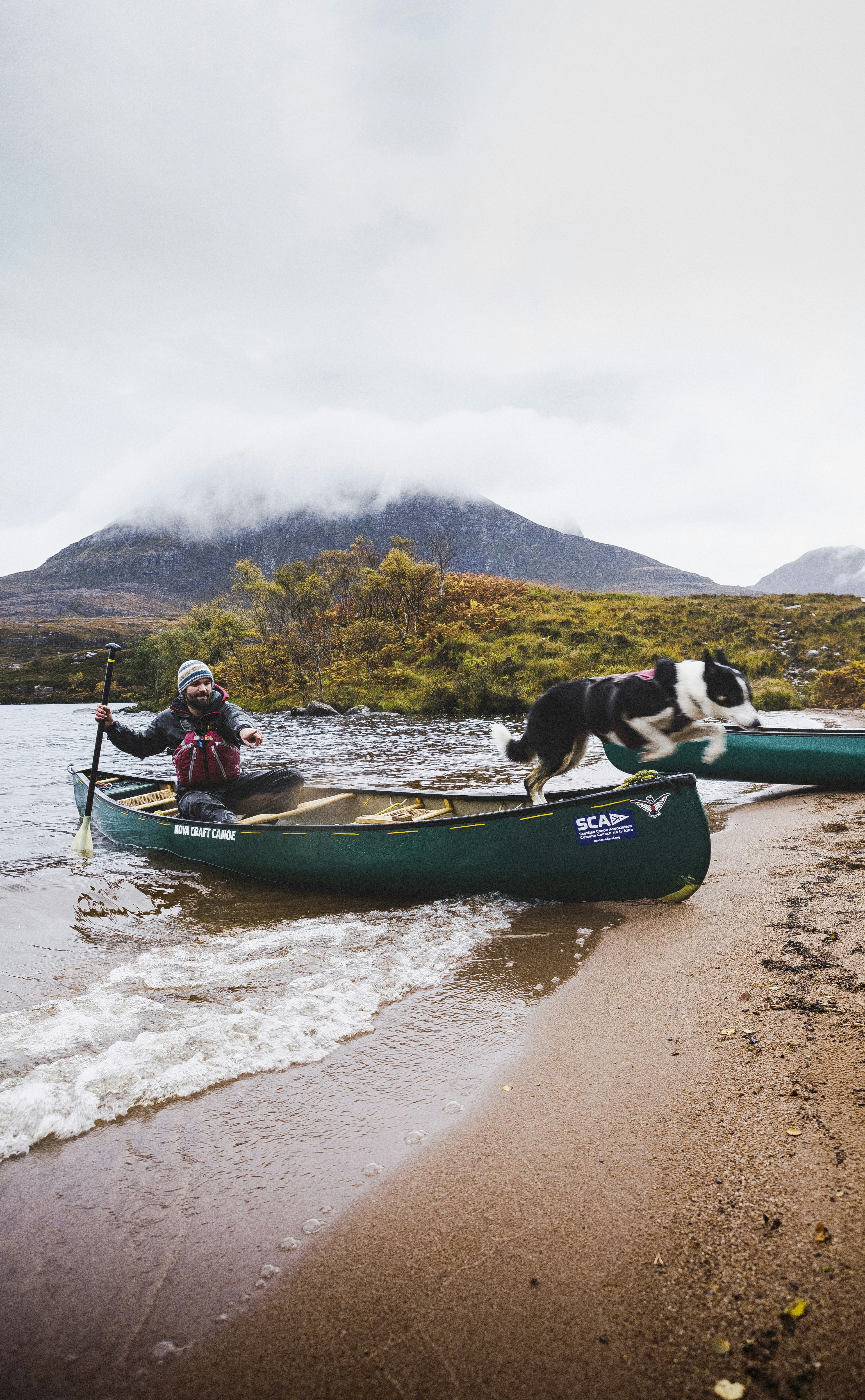 A border collie leaps out of a canoe as its guide beaches a canoe on the shore of Loch Lurgainn, under the peak of Stac Pollaidh.