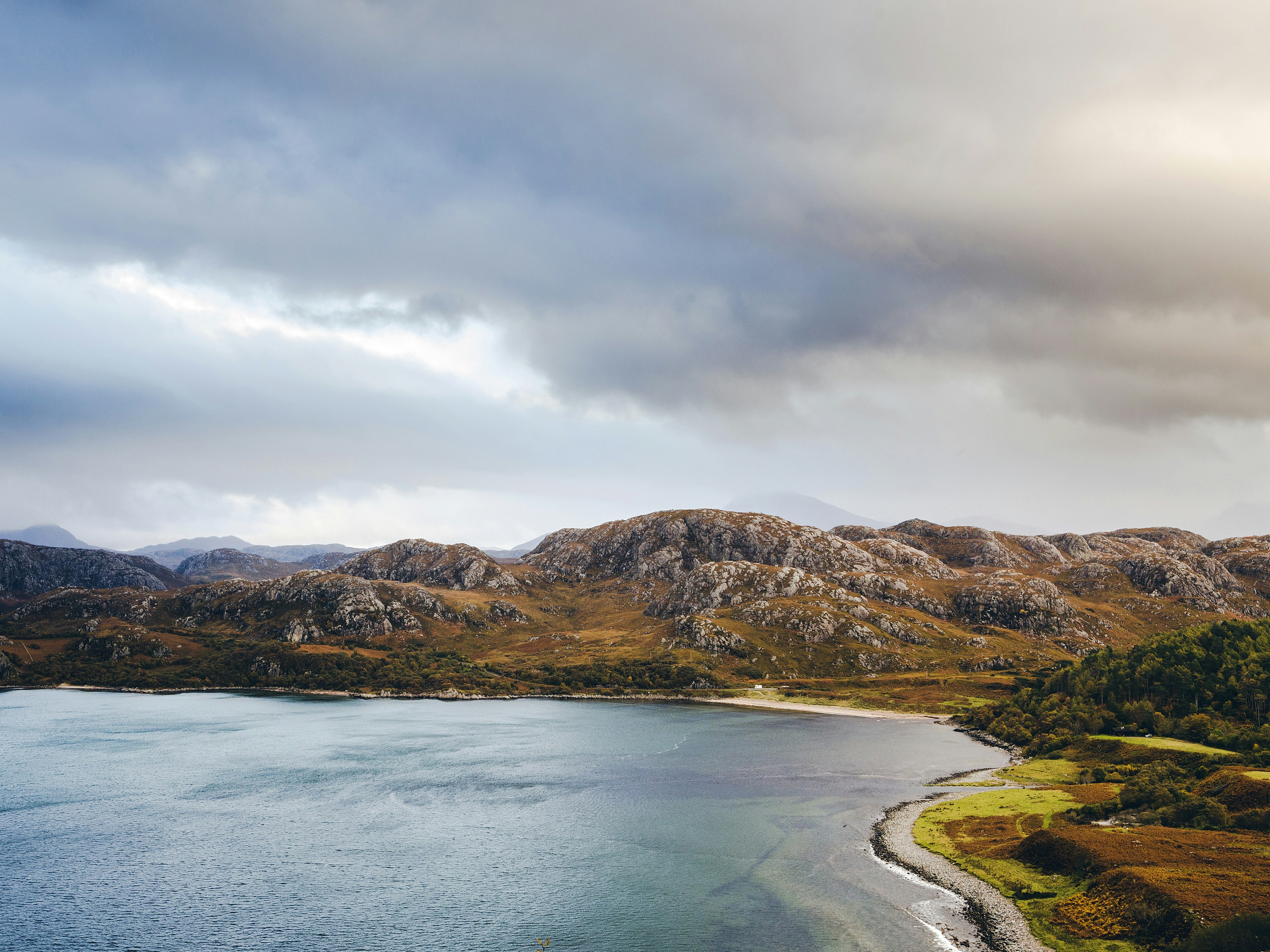 One of the westernmost stretches of the NC500 passes by a small beach at Gruinard Bay. An image of the bay with trees on the right and mountains in the background, under a cloudy sky.