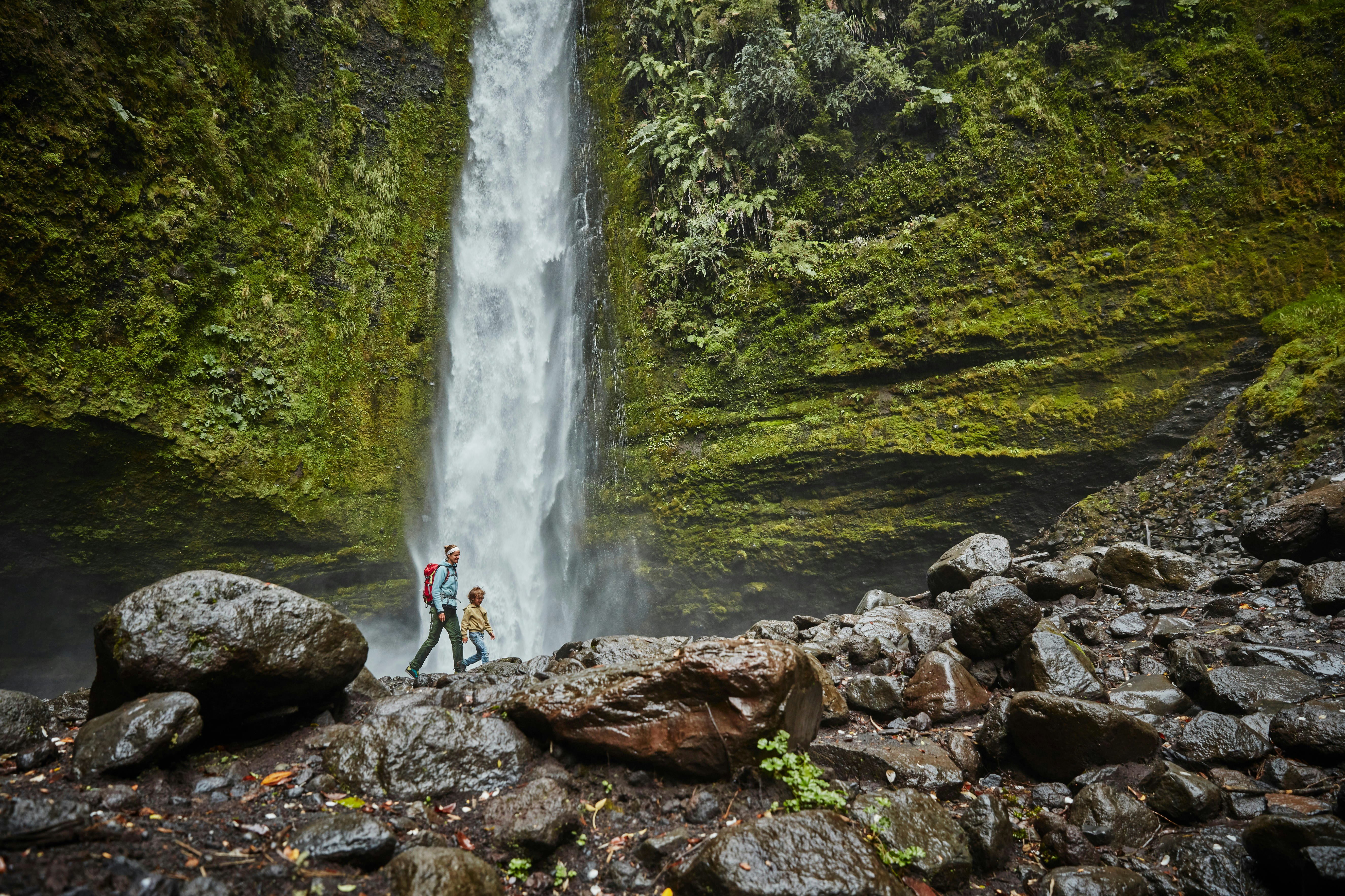 A mother and son walking by the Las Cascadas waterfall, which plunges down a mossy rock face onto wet boulders below