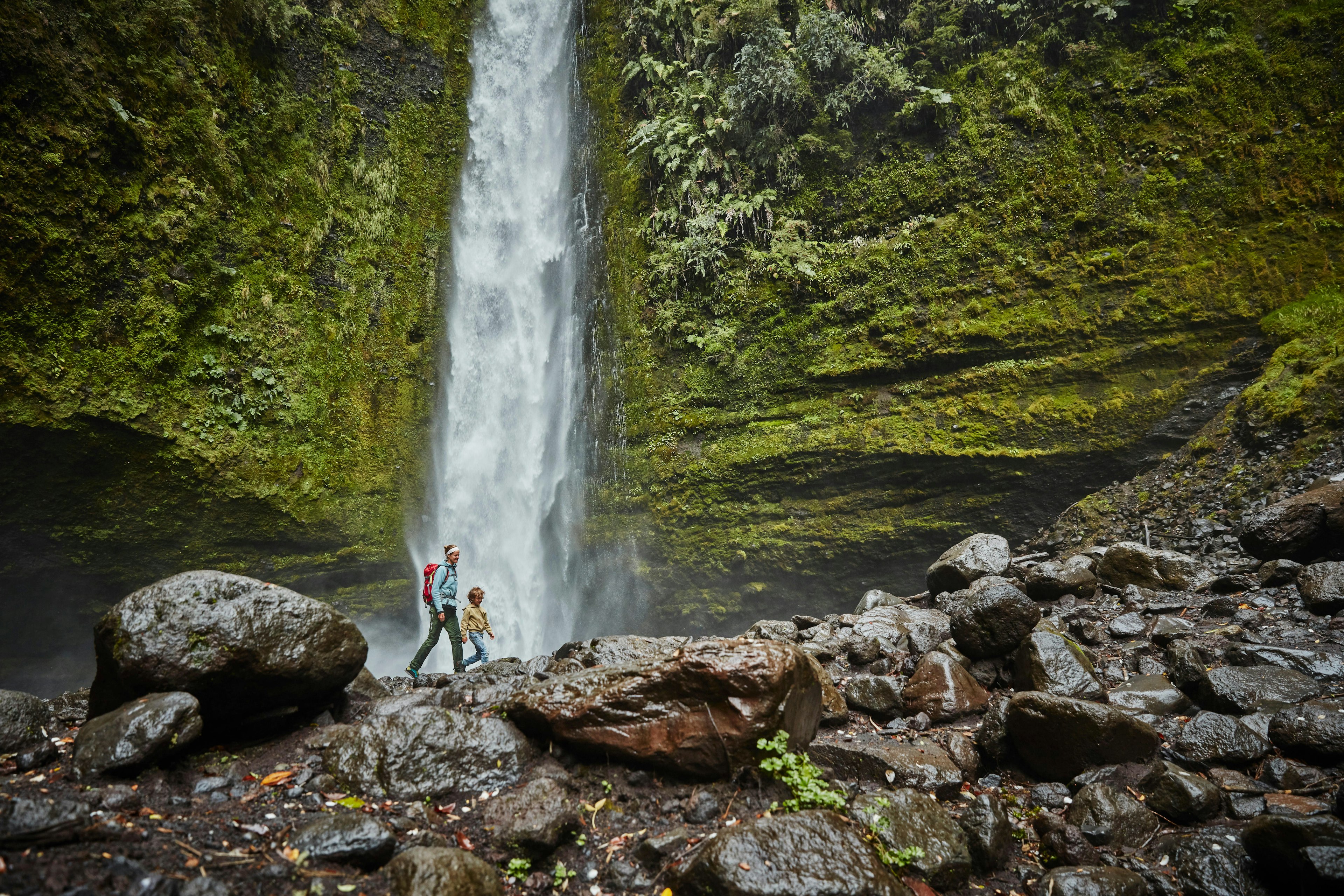 A mother and son walking by the Las Cascadas waterfall, which plunges down a mossy rock face onto wet boulders below