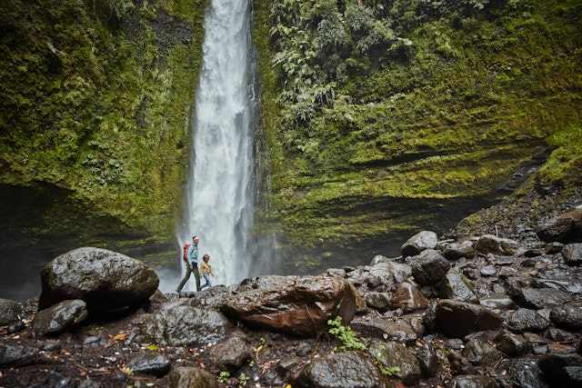 A mother and son walking by the Las Cascadas waterfall, which plunges down a mossy rock face onto wet boulders below