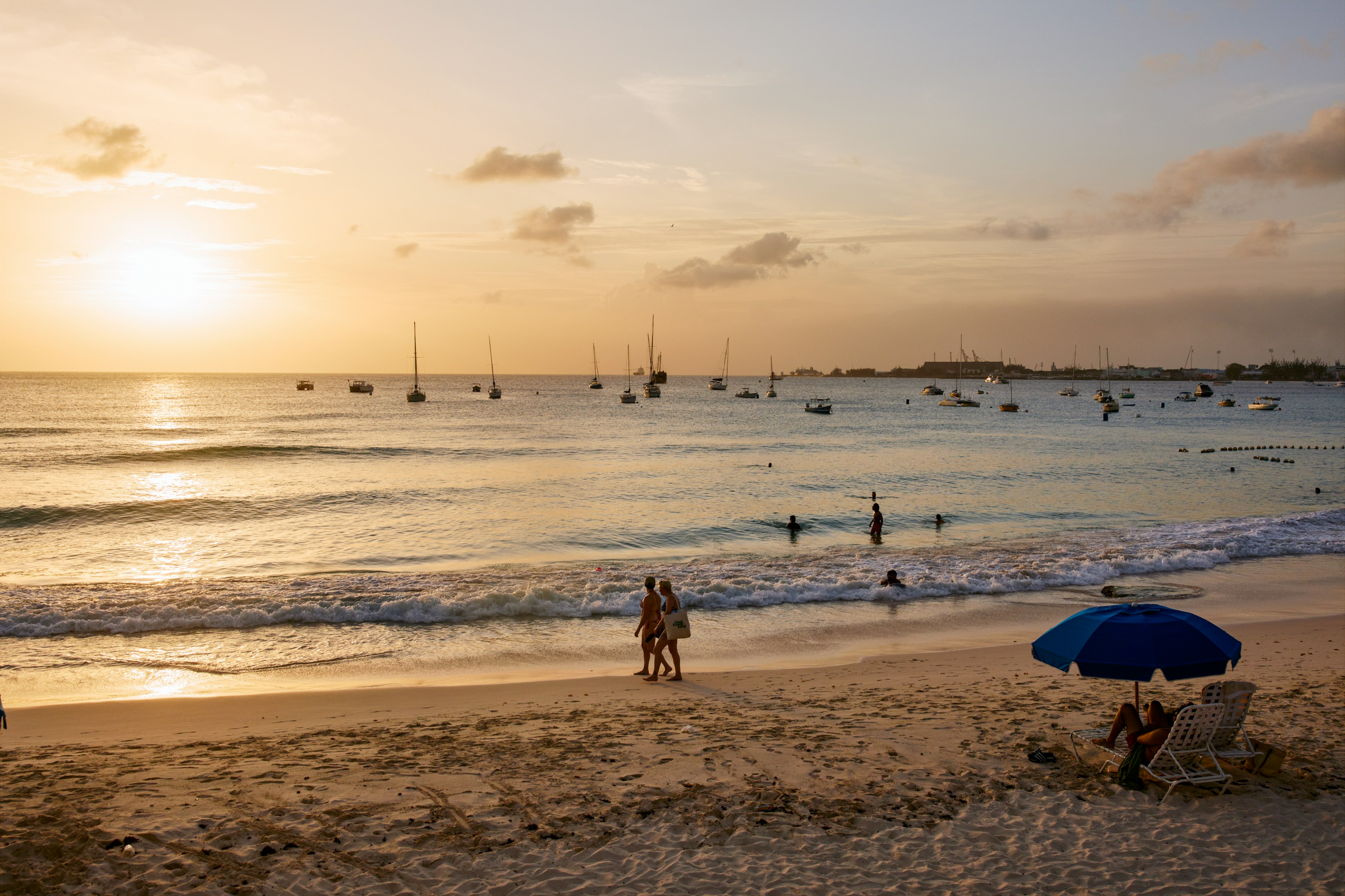 A couple walks along the beach at Pebbles Beach at sunset, with the masts of moored sailboats in the distance, Barbados