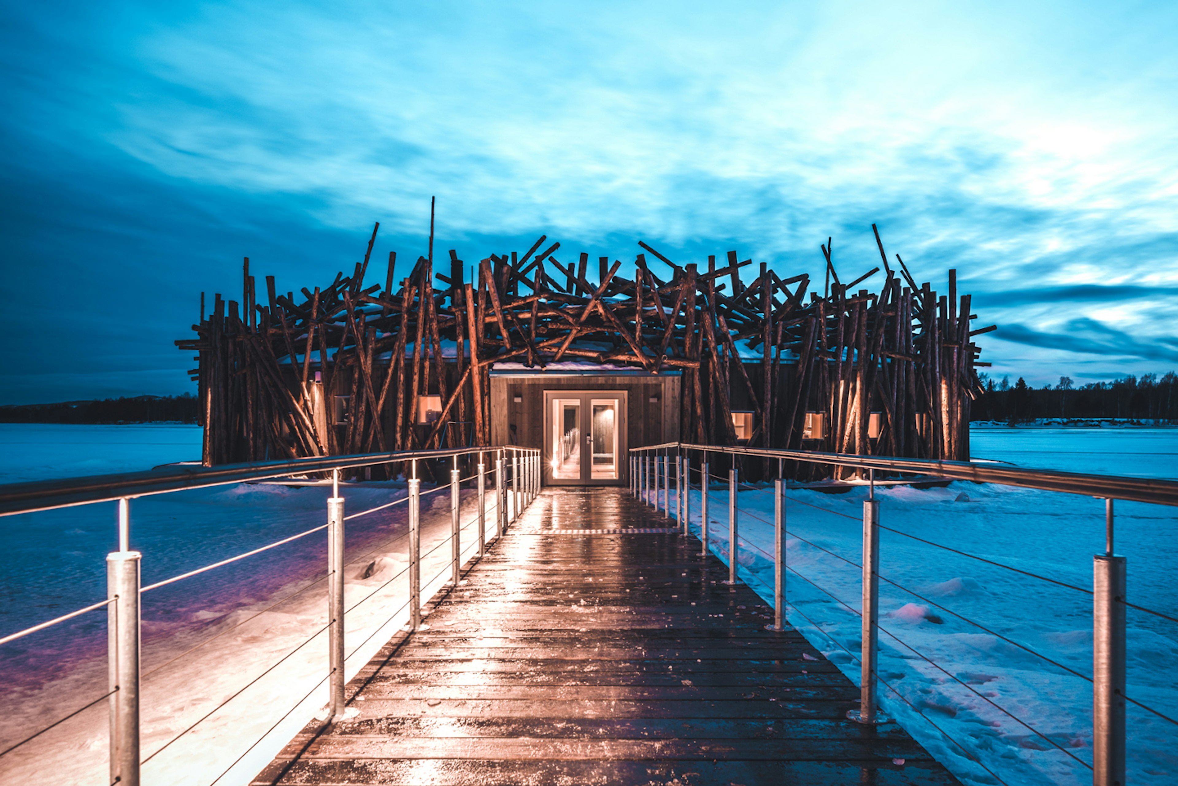 A wooden walkway through a frozen lake leads to a large circular wooden structure that houses a floating spa