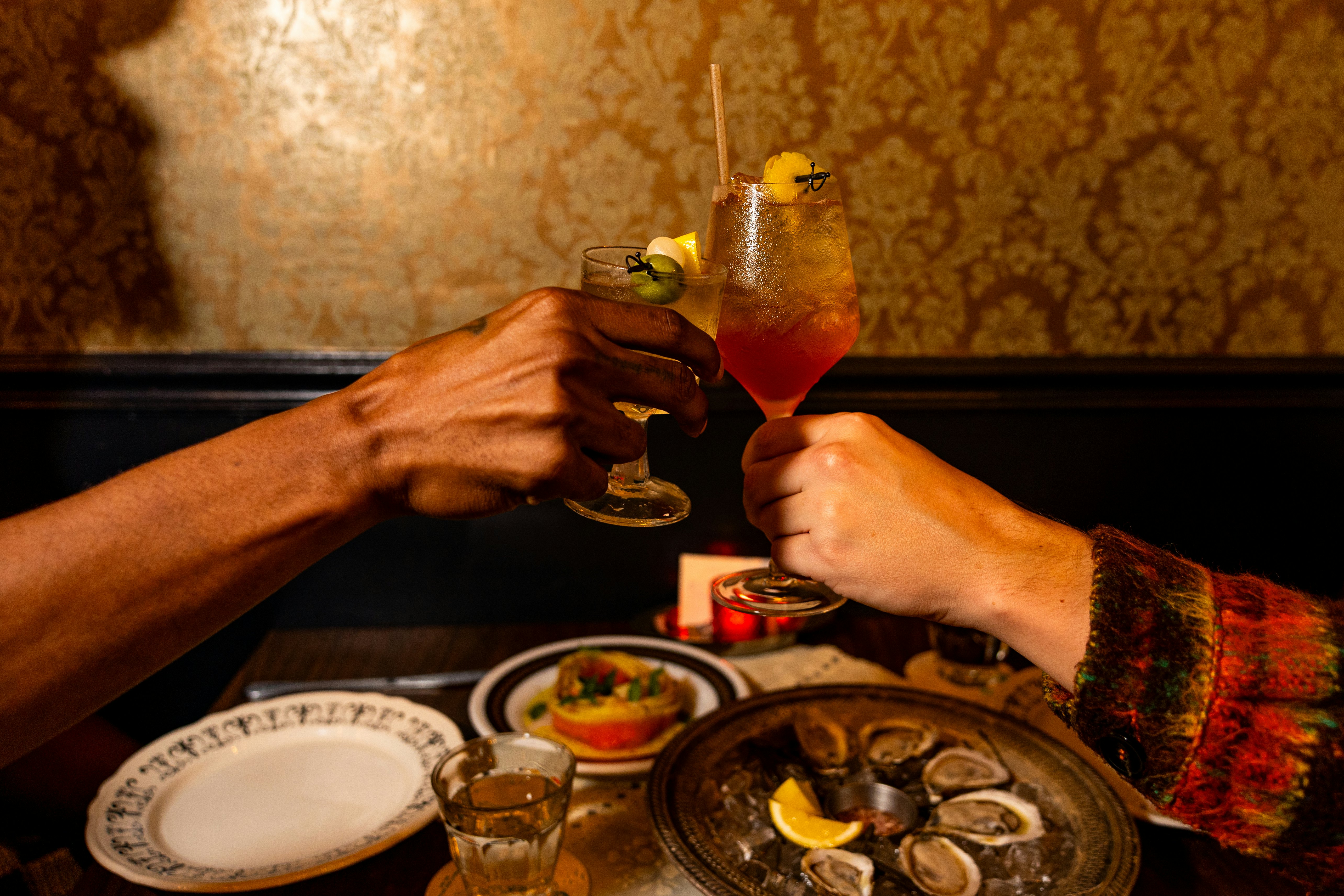 Two people toast their cocktails at FET FISK restaurant in Pittsburgh. A plate of raw oysters is between them.