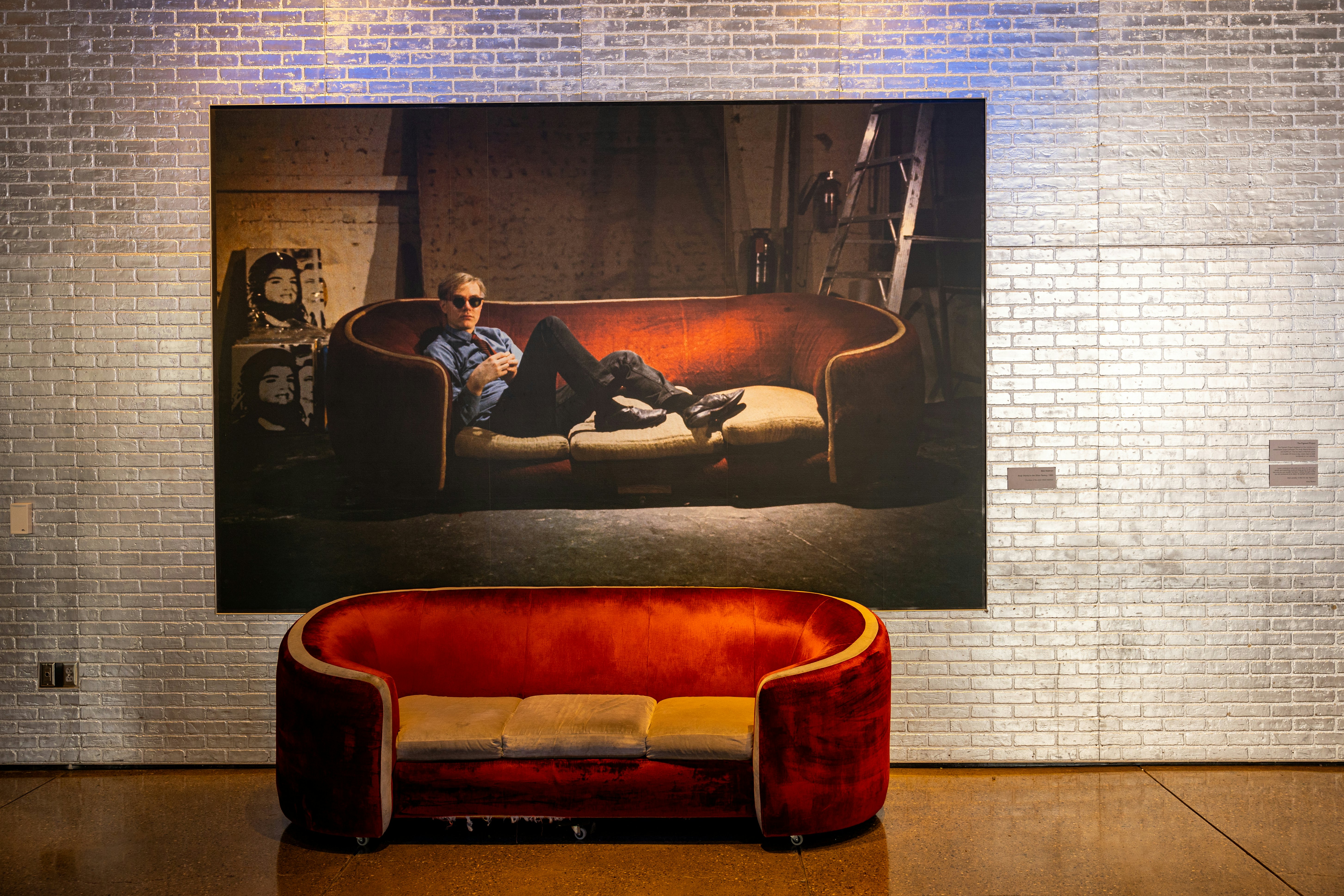 A red sofa in front of a painting of Andy Warhol reclining on the same sofa