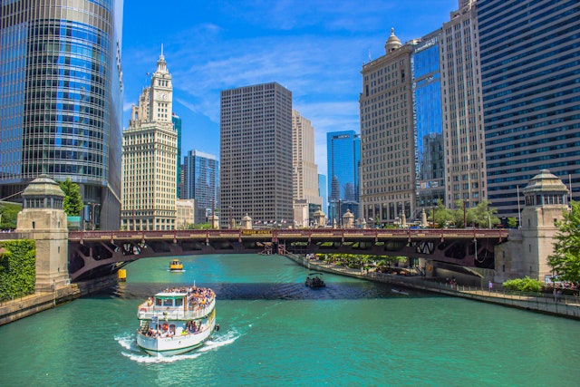 Boats loaded with tourists cruise along a river that flows between tall skyscrapers in a high-rise city