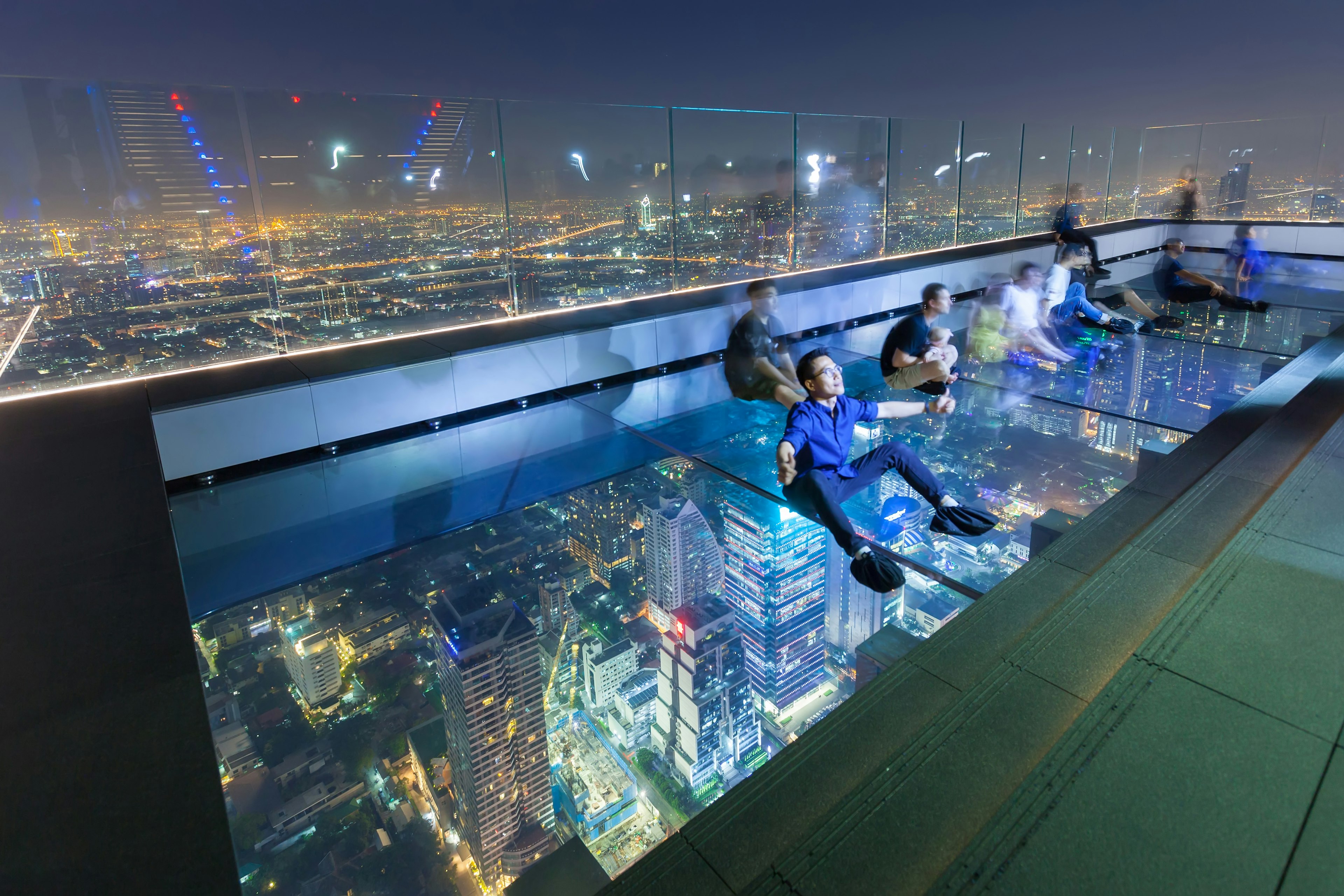 Visitors sit and lie on the glass floor at night at the SkyWalk rooftop of the King Power Mahanakhon building in Bangkok as the lights of the city skyline, some way below, glitter.