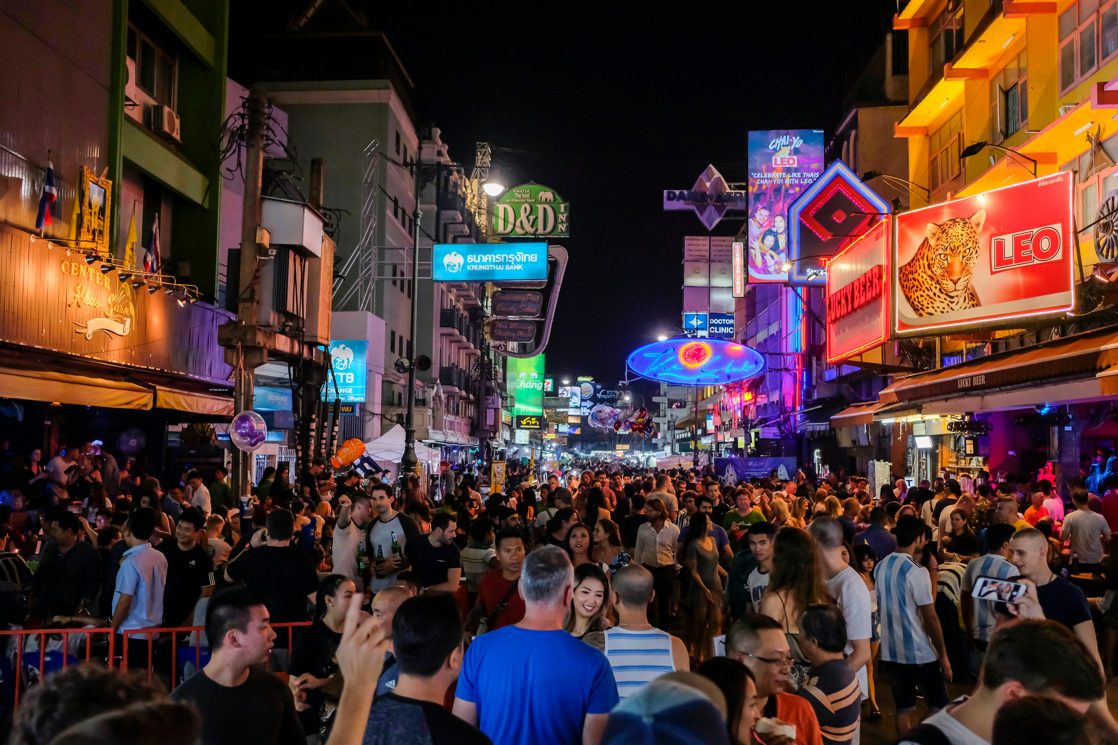 Hundreds of people walk at night along Khao San Road in Bangkok, with neon signs and billboards overhead. This area is very popular with backpackers and other travelers.