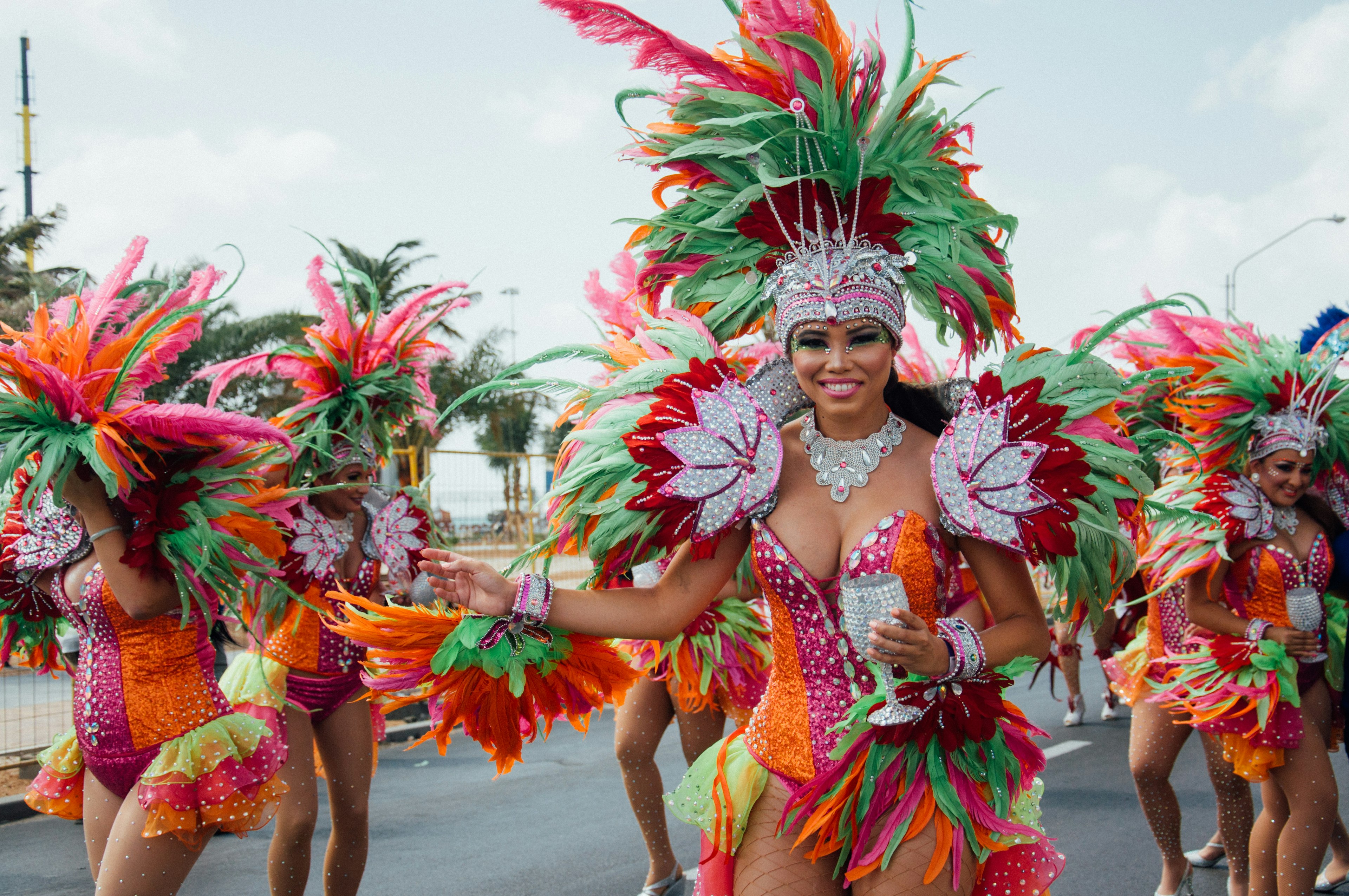 A woman in a bright orange green short dress and a big decorative crown with feathers walking and smiling during the Carnival procession, Aruba