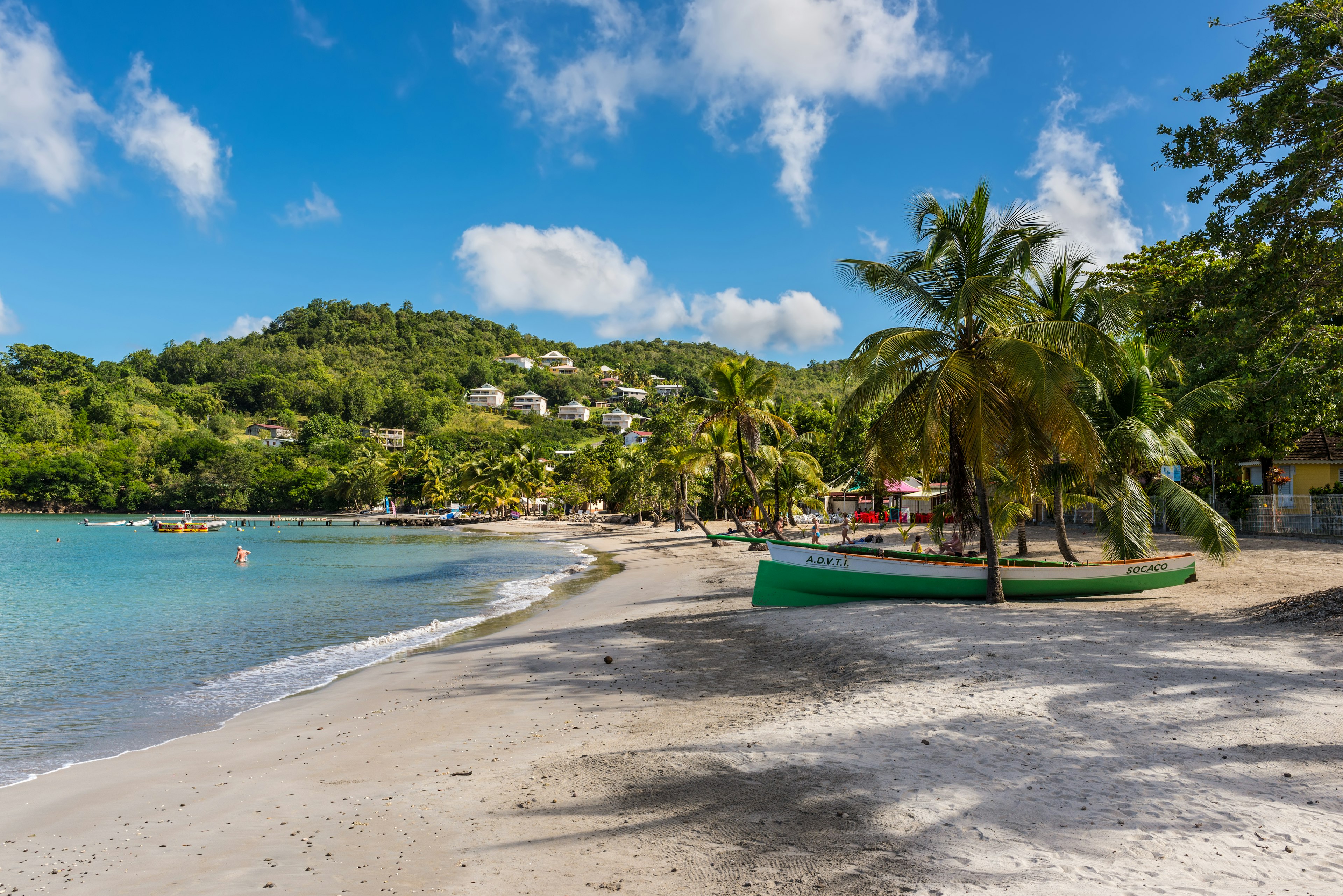 A green boat sits on a a beach surrounded by palm trees with villas in the hills beyond, Les Trois-Îlets, Martinique