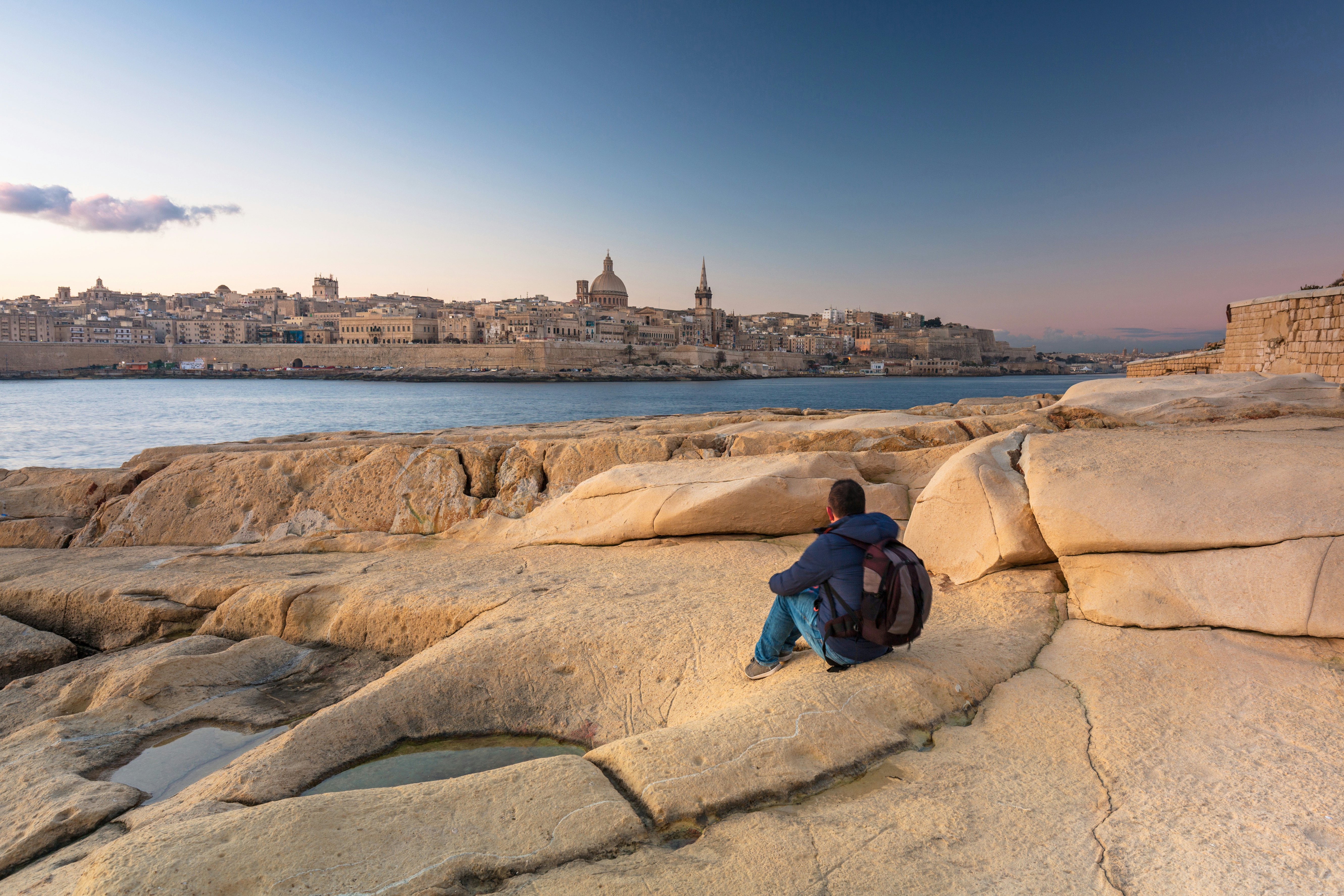 Man sitting on the rock and watch beautiful architecture of the Valletta city at dawn.