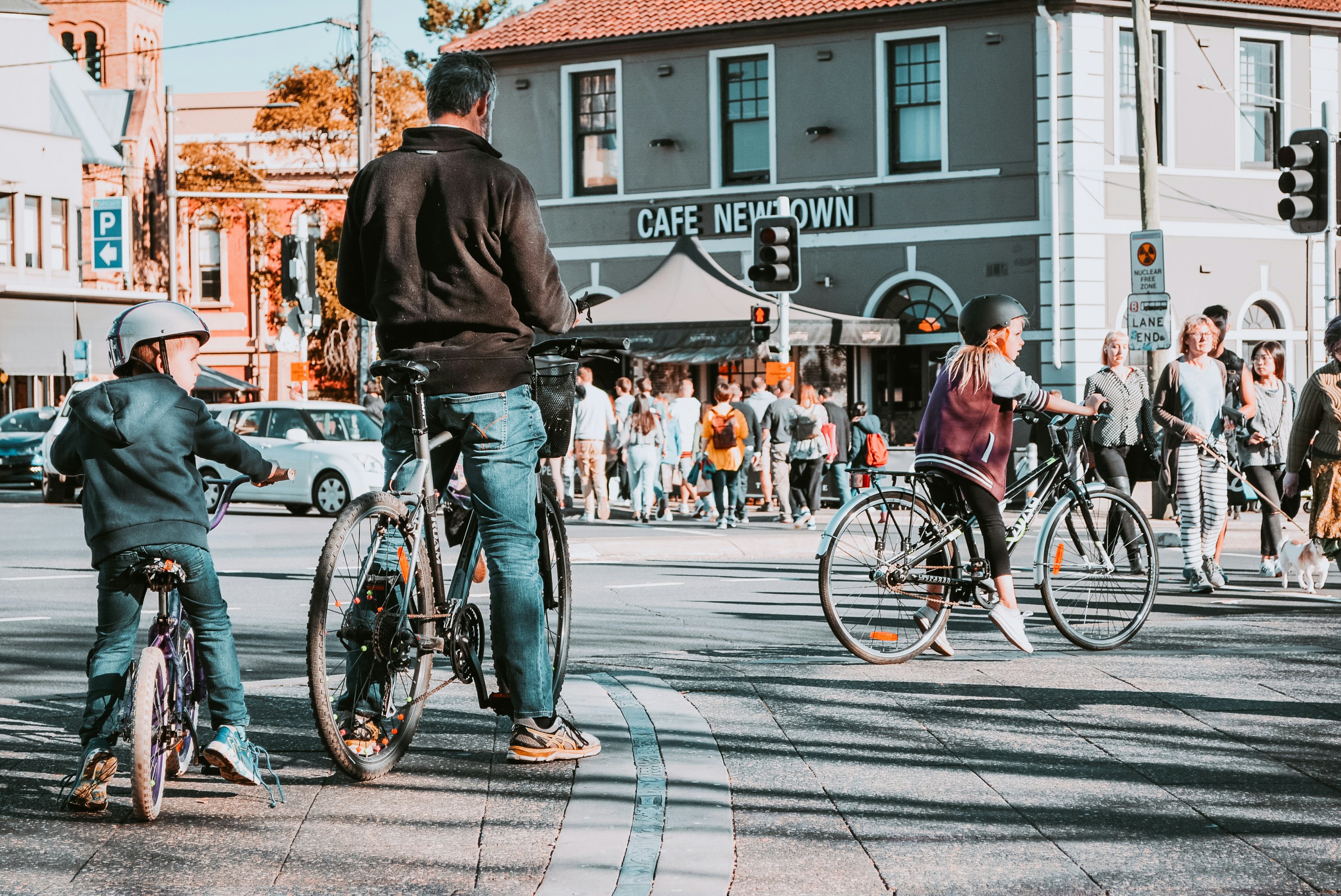 An adult and two child cyclists stop on a street in busy city neighborhood. Pedestrians line the sidewalk and crosswalk.