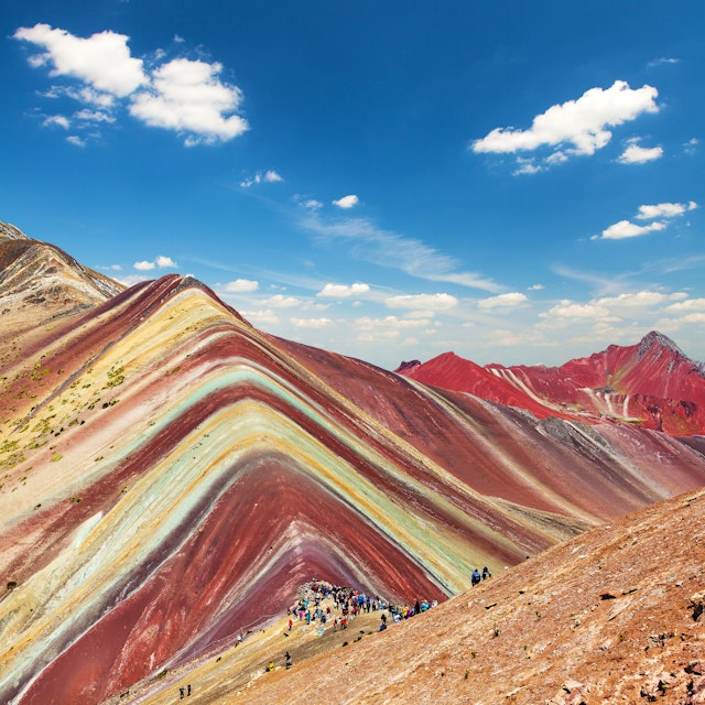 A wide-angle shot of Rainbow Mountain near Cuzco in Peru. The mountain's striations are brightly coloured, and a group of hikers has gathered to admire it.