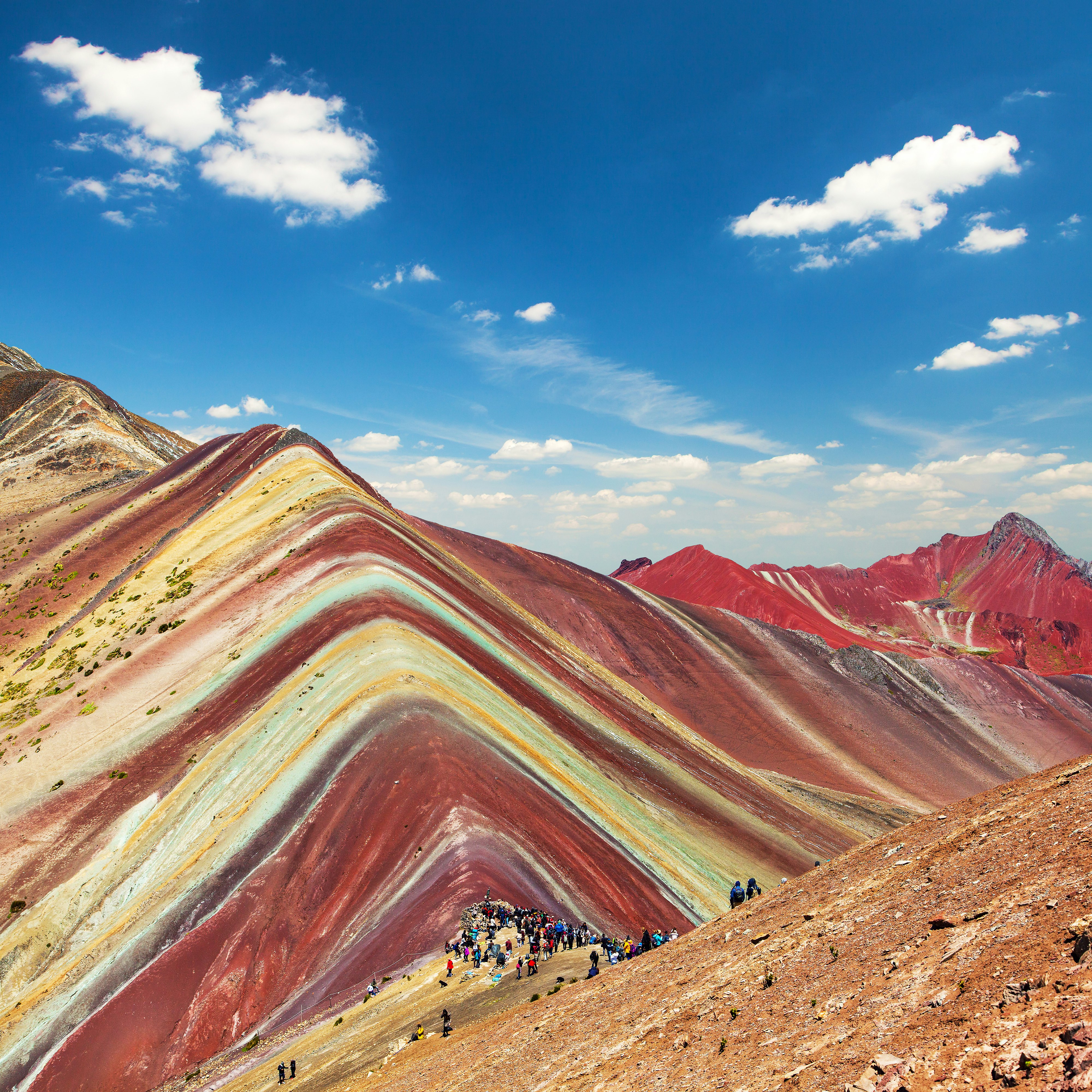 A wide-angle shot of Rainbow Mountain near Cuzco in Peru. The mountain's striations are brightly coloured, and a group of hikers has gathered to admire it.