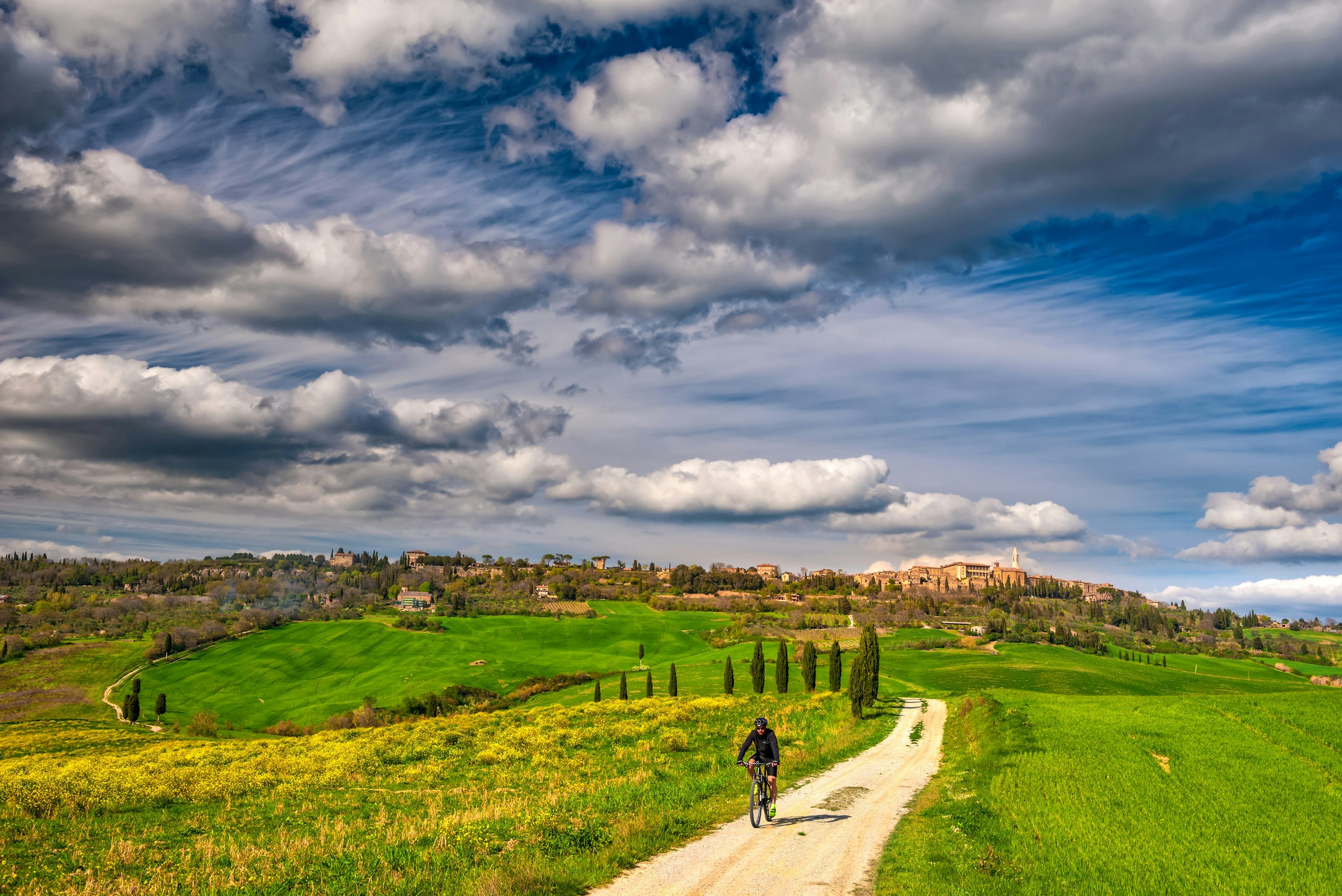 A cyclist pedals along a winding path through countryside with a small town on a distant hilltop