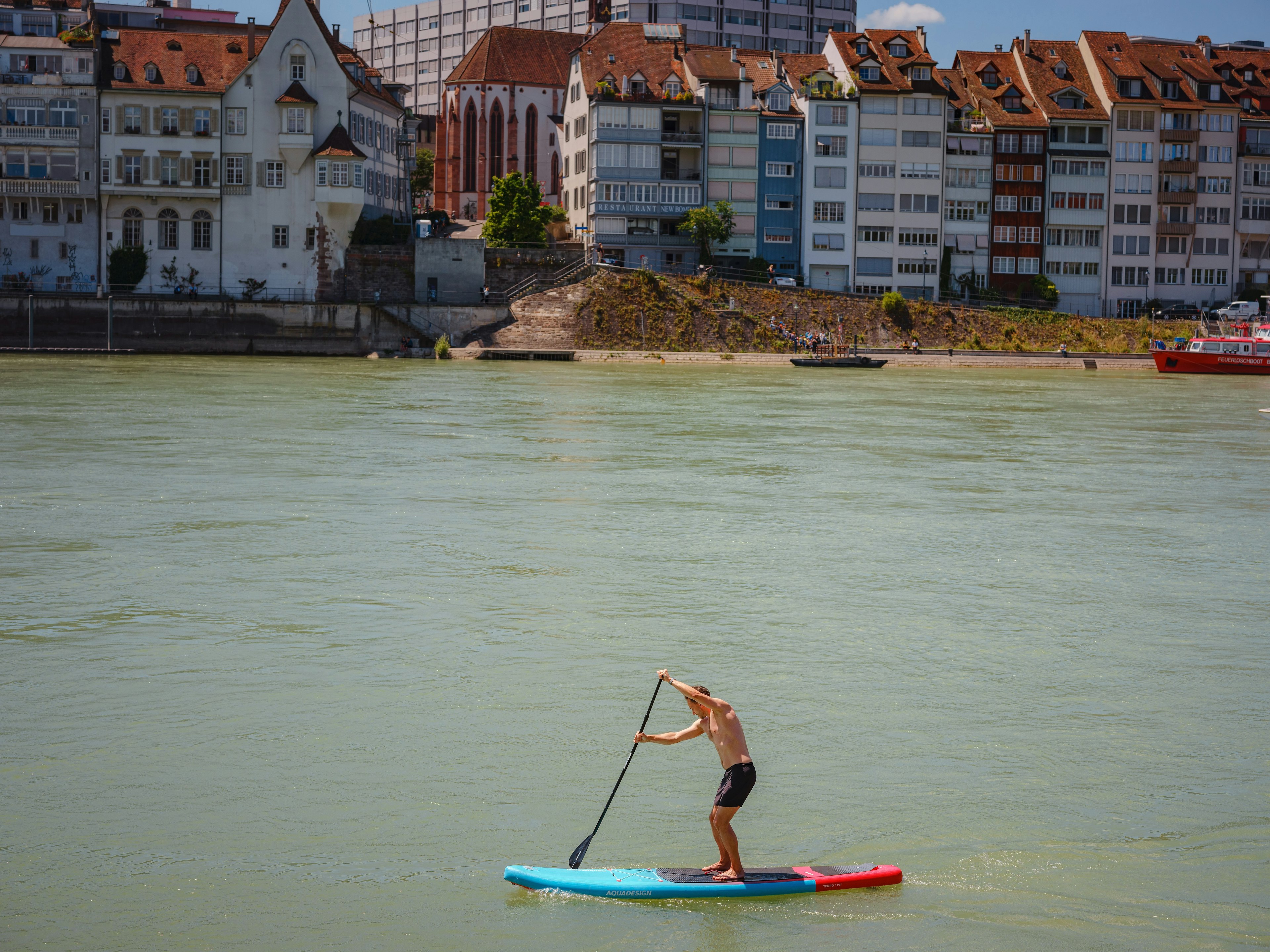 A man on a stand-up board floats on the river Rhine, in the city center of Bern, with apartment buildings along the shoreline