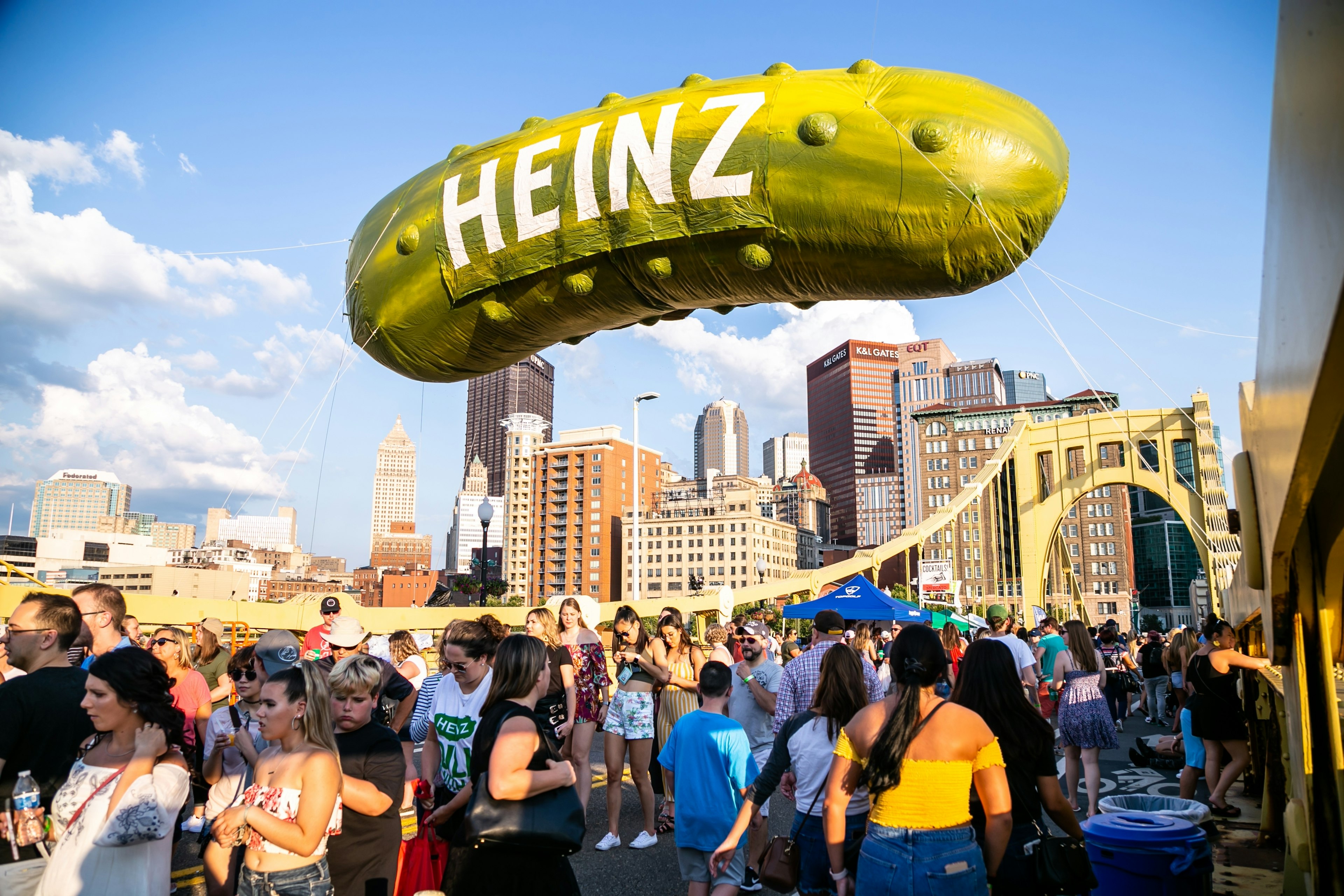 A giant balloon in the shape of a pickle, with large white letters spelling HEINZ, is tethered to a bridge in Pittsburgh while a large crowd mingles on the bridge itself