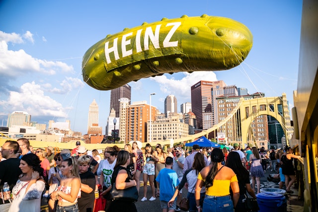 A giant balloon in the shape of a pickle, with large white letters spelling HEINZ, is tethered to a bridge in Pittsburgh while a large crowd mingles on the bridge itself