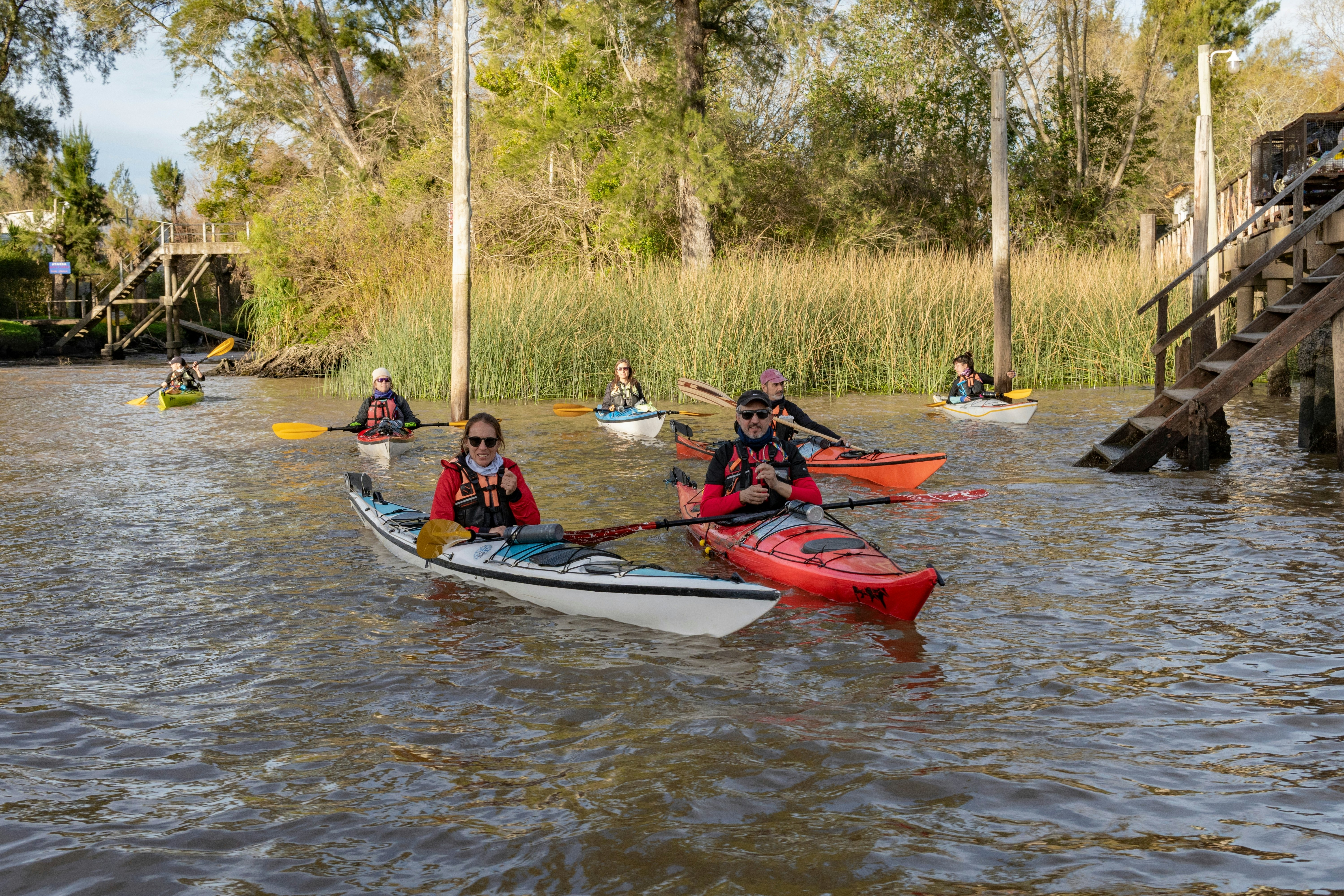 A group of people in kayaks on the Sarmiento River, part of the Paraná Delta, Tigre, Argentina. Wooden steps leading to the water are pictured, along with tall aquatic grass.