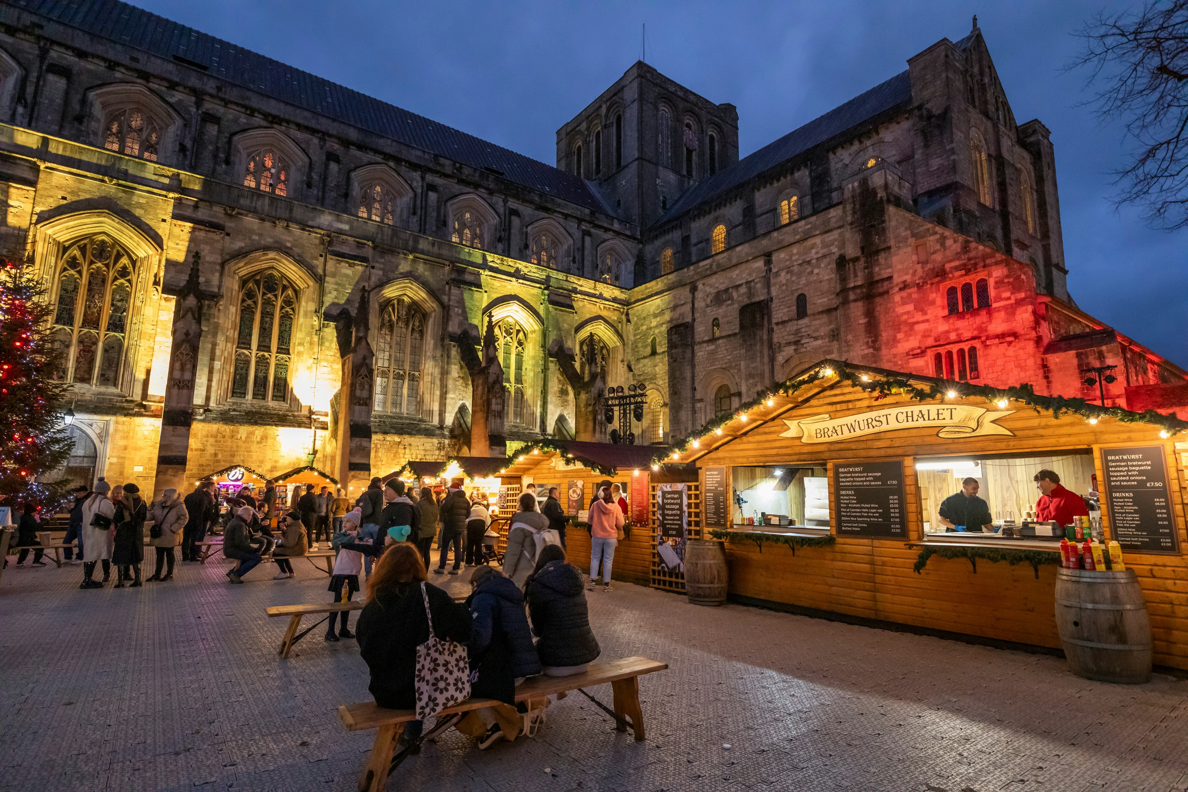 Wooden huts and picnic benches around a large cathedral lit up during the Christmas season