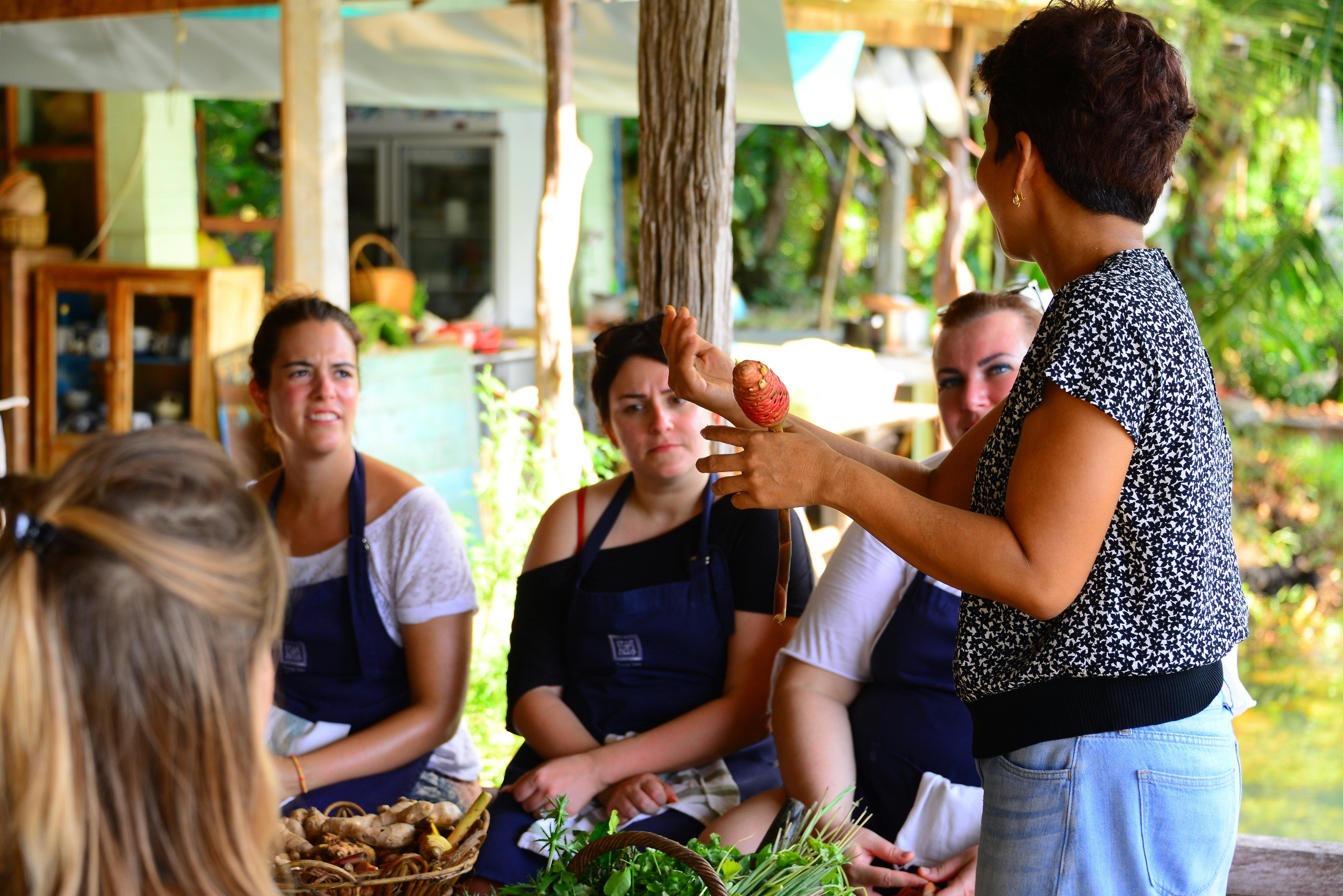 A woman holding a vegetable in front of a table with baskets of produce leads a class on Thai cooking as she talks to several other participants wearing aprons