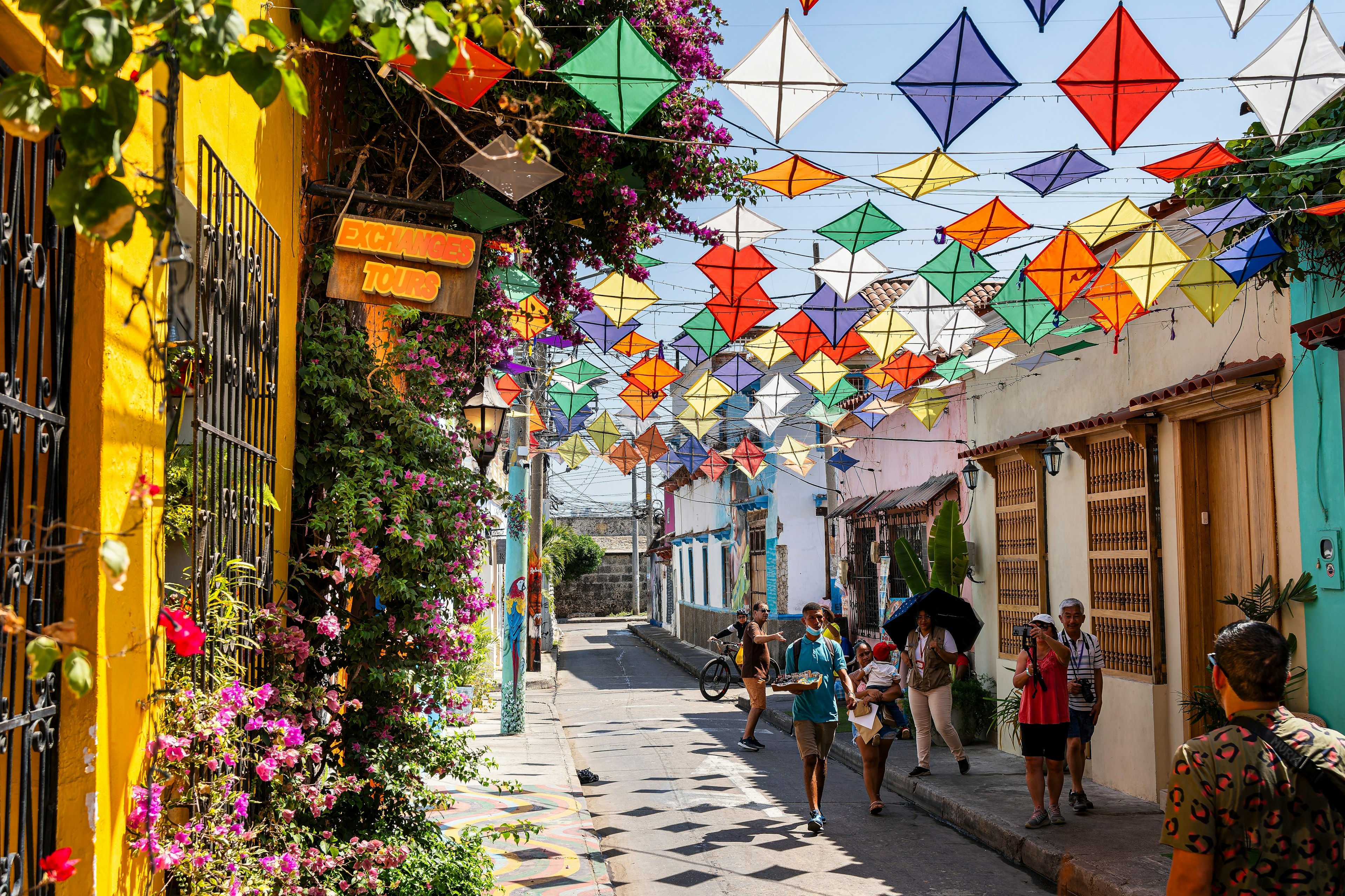 People walk down an alley with colorful paper kits strung above head height and bougainvillea growing by brightly painted walls