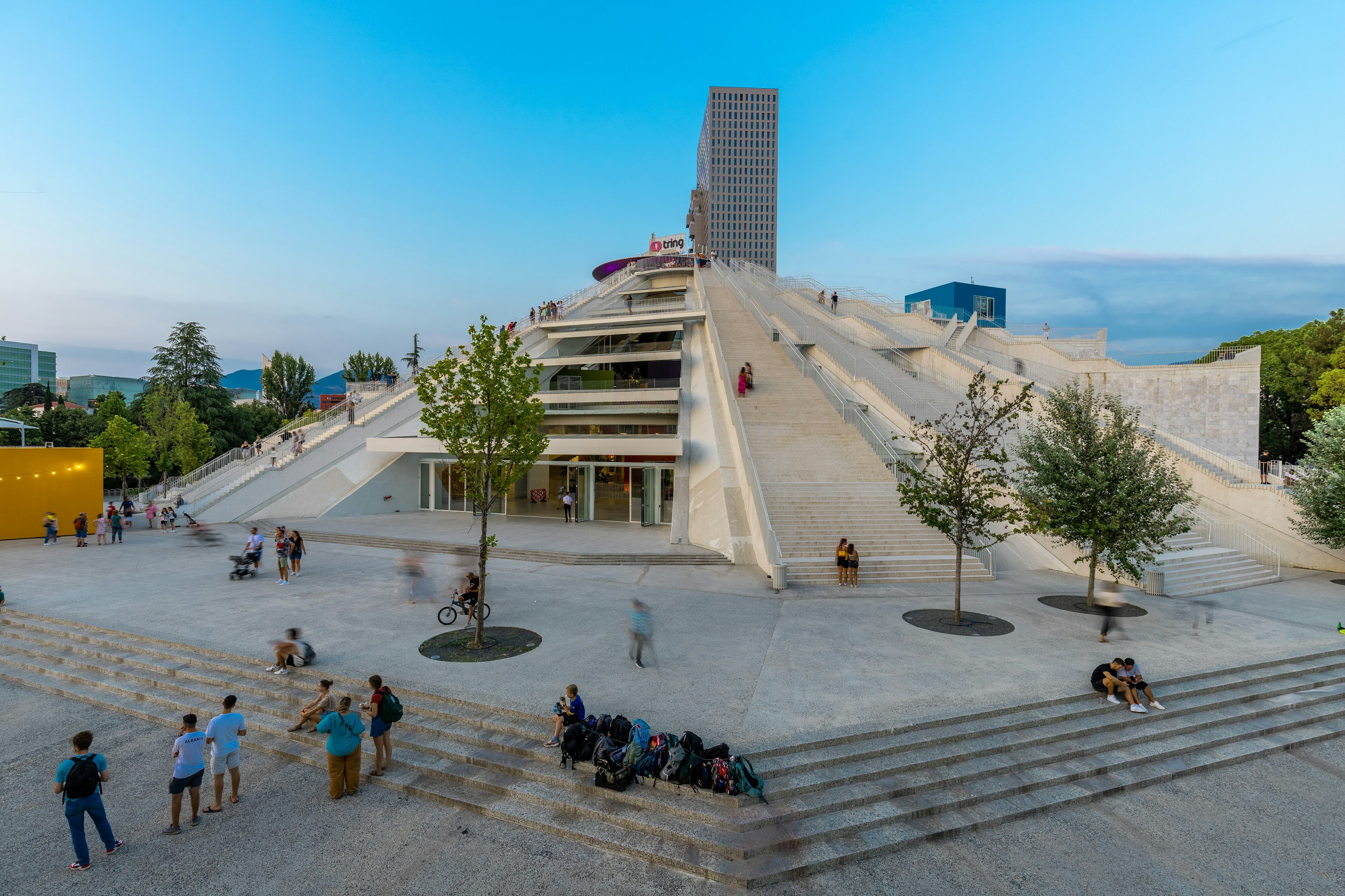 People mill around the base of a pyramid-shaped building with some climbing up the side at dusk