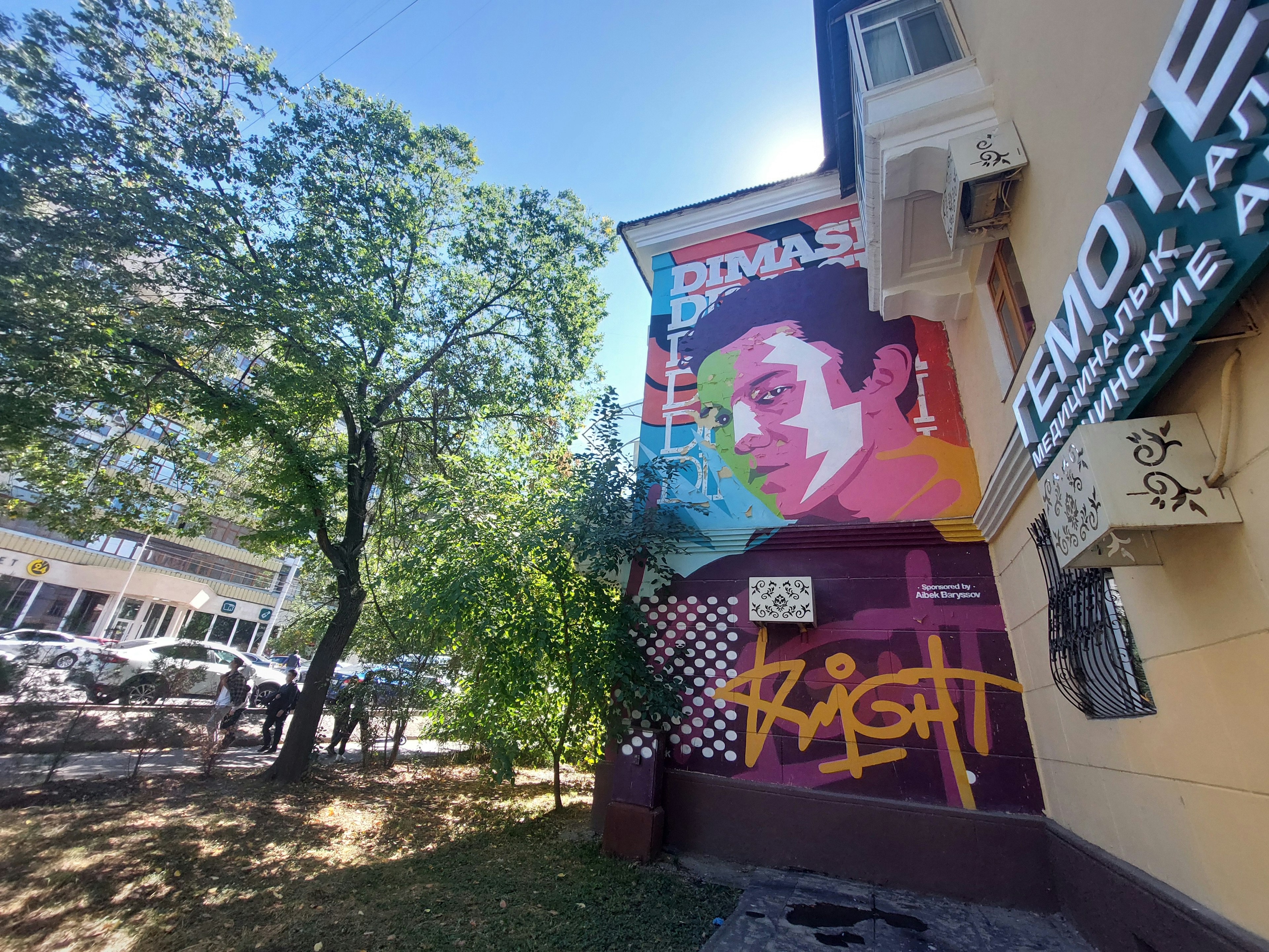 A mural depicting the singer Dimash on a street with trees with cars in the background in Almaty, Kazakhstan