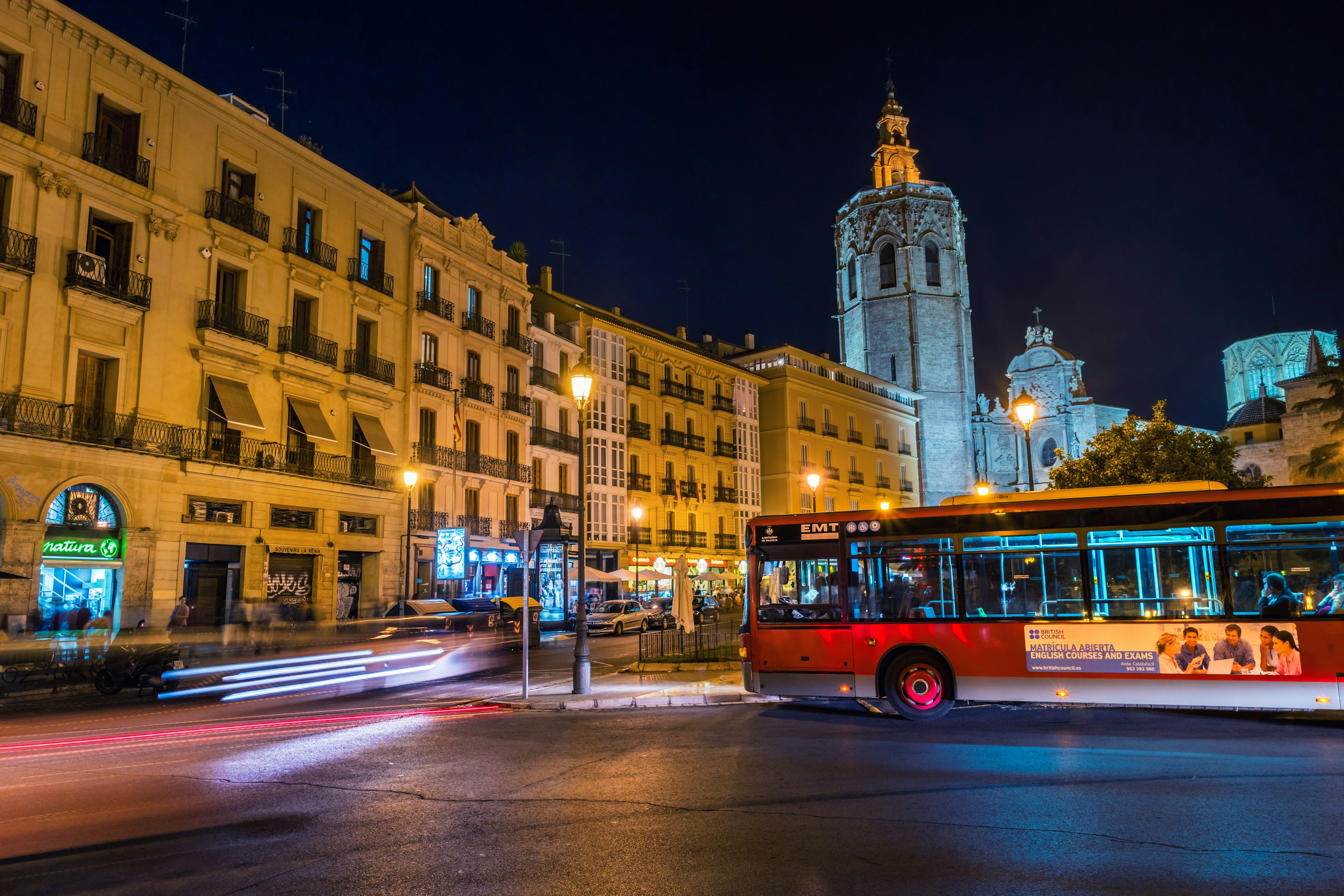 A red bus drives past illuminated historical buildings at night at Plaça Reina, central Valencia, spain