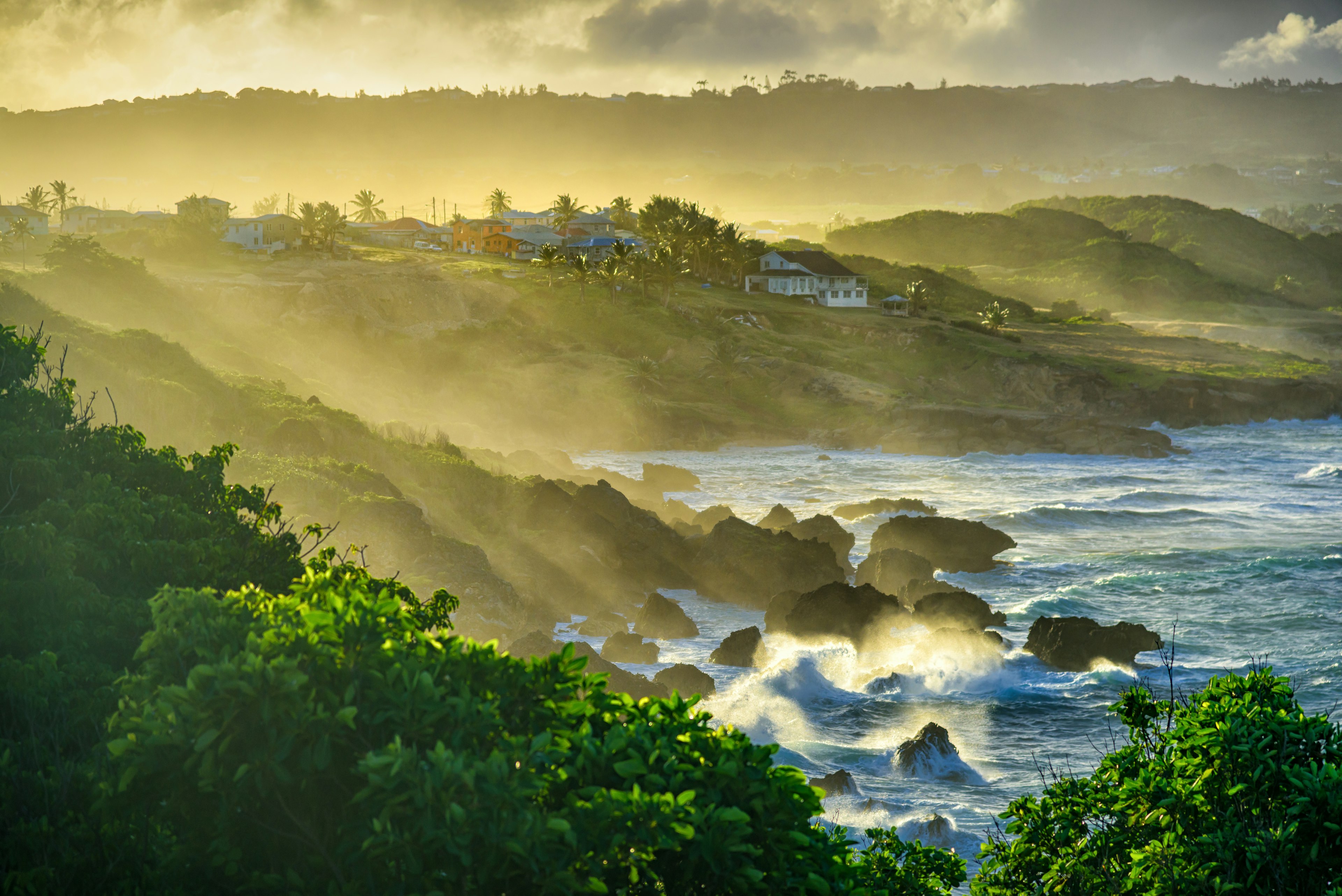 Beams of sun illuminate the water spray hitting the land after strong waves splash on the rock at sunset, Ragged Point, Barbados