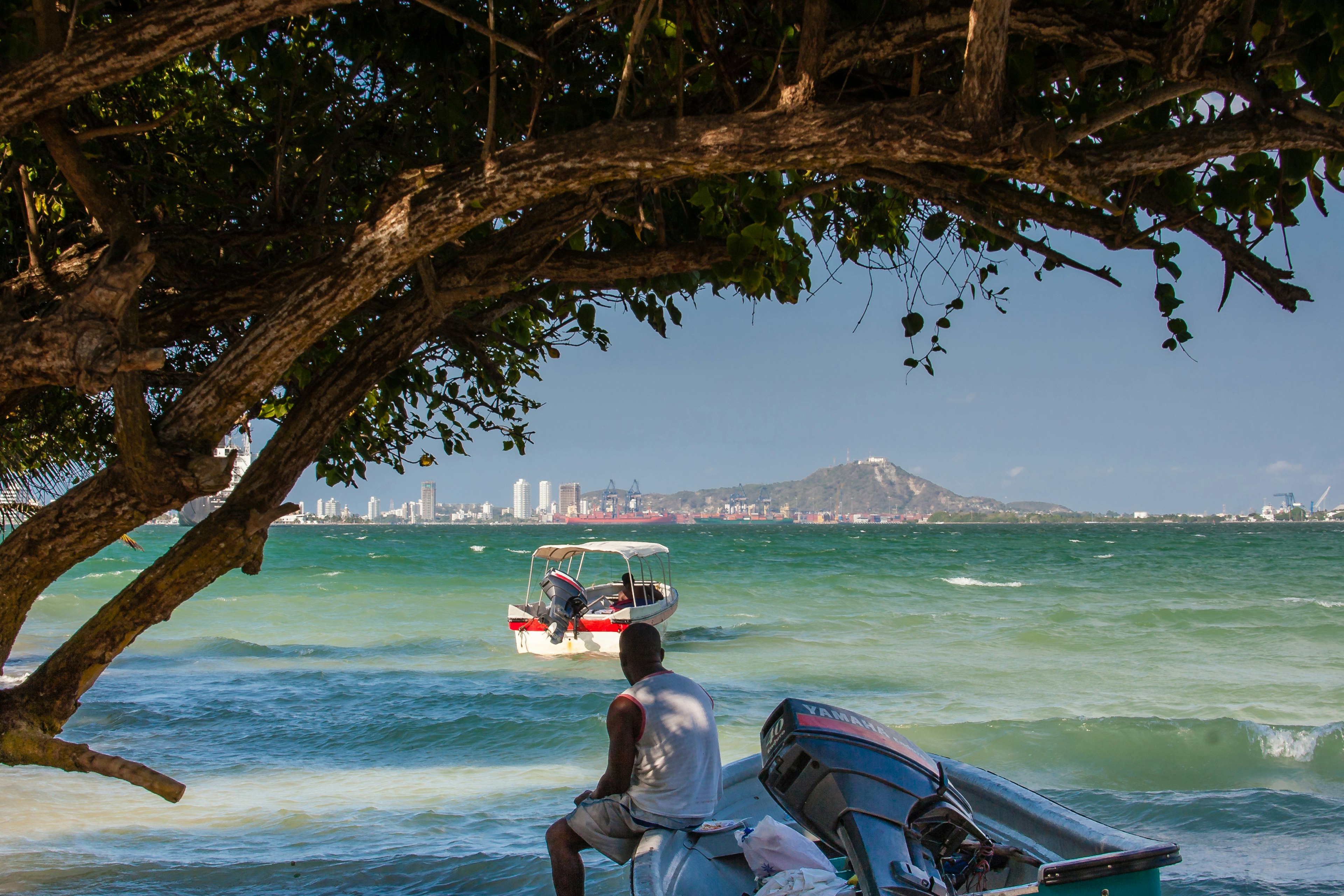 A mean sits in a boat on shore, looking out at another boat on the water and the city skyline and a mountain in the distance, Tierra Bomba, Cartagena, Colombia