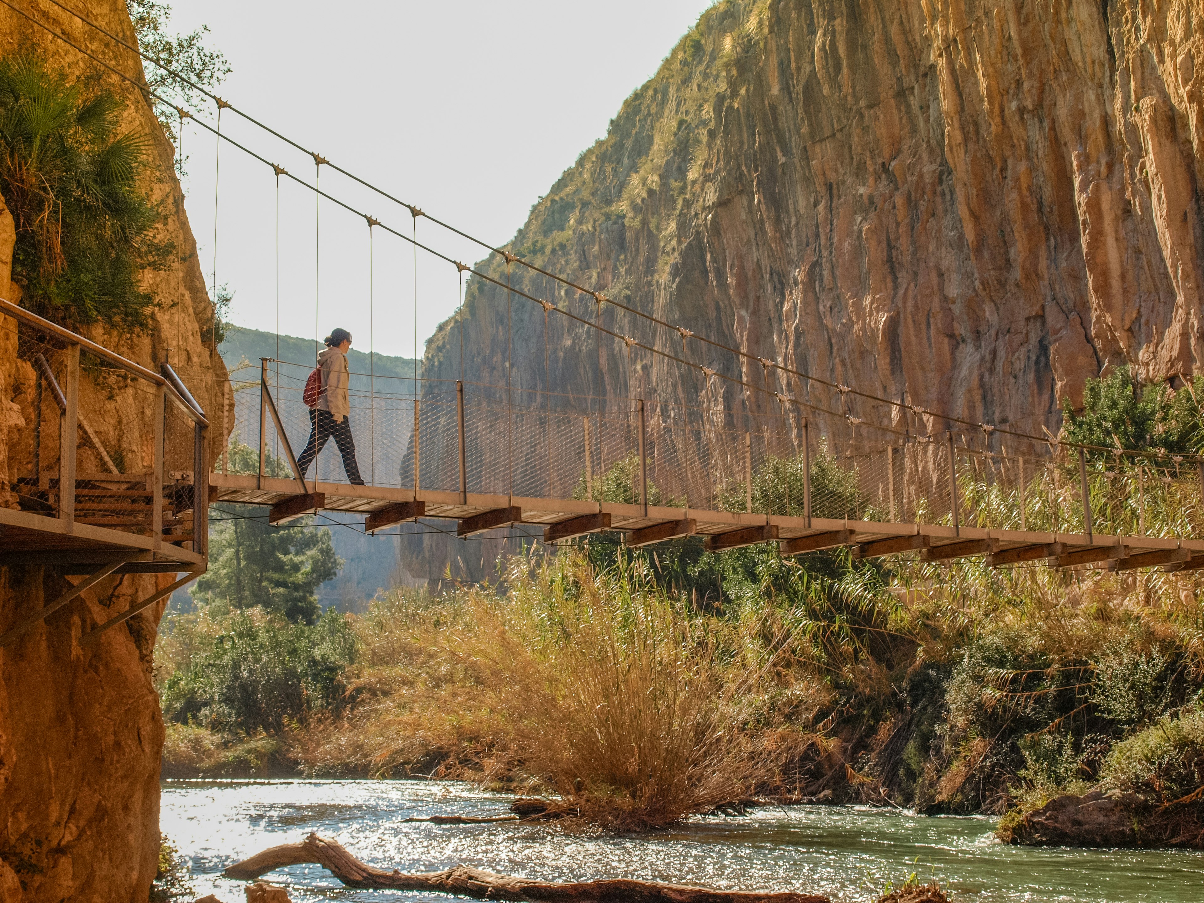 A hiker walks over a suspension bridge above a river, linking up two sides of a gorge